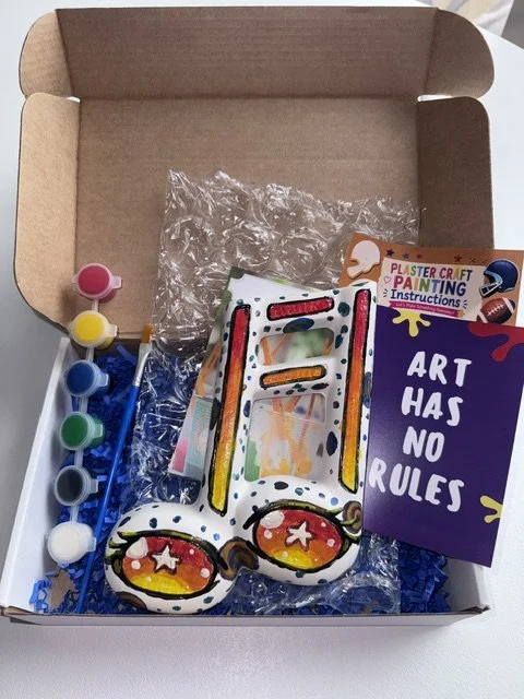 A colorful fish-shaped plush toy, a white fish-shaped ceramic craft, and a paper sign that reads 'Ceramics on Wheels, Arts & Crafts, We come to you.' The setup includes various small bottles of paint in red, black, yellow, white, orange, and green, along with two paintbrushes.