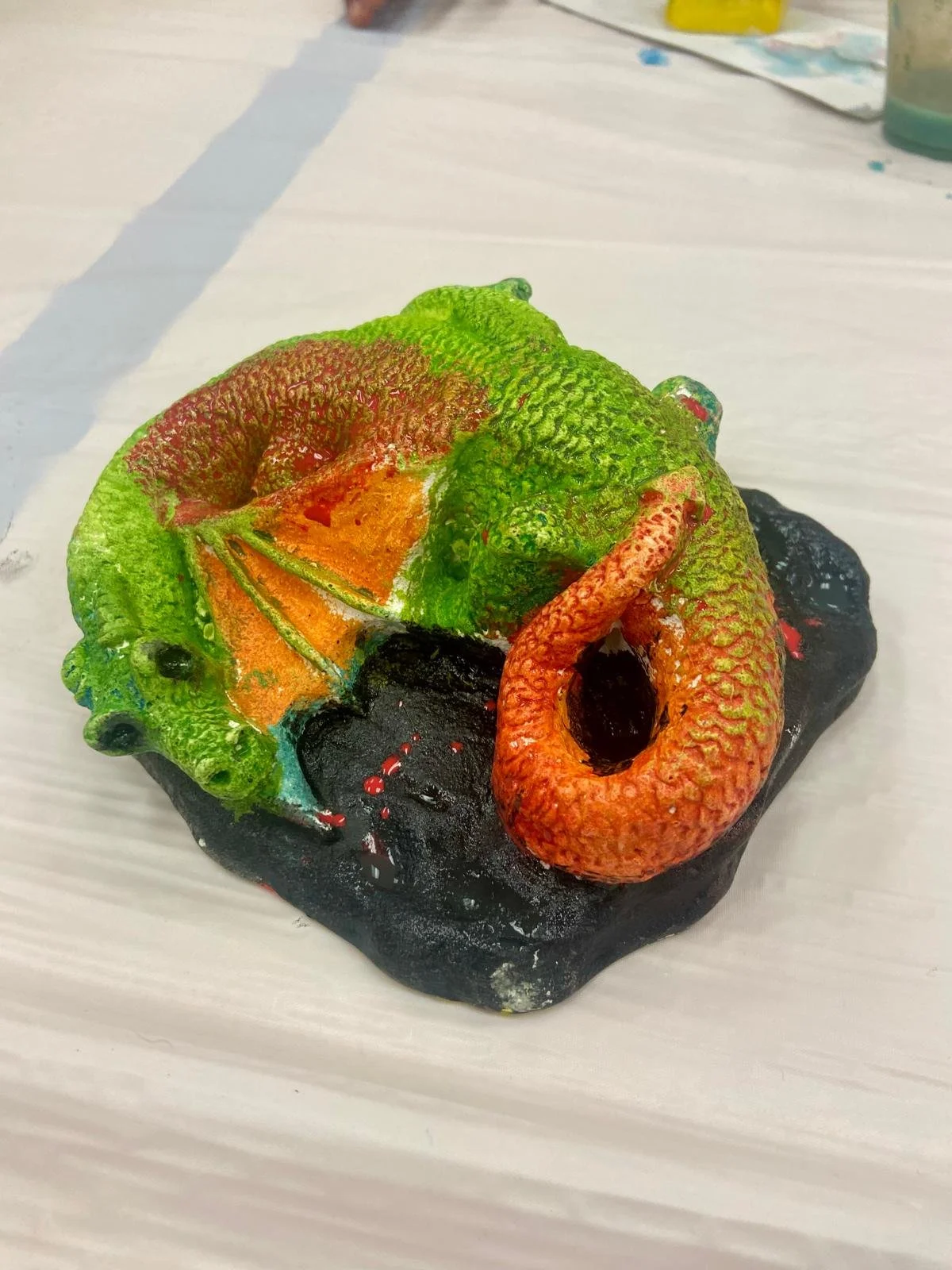 A multicolored, realistic model of a chameleon lizard on a black base, with textured skin and exaggerated features, made of a sculpting or modeling material.