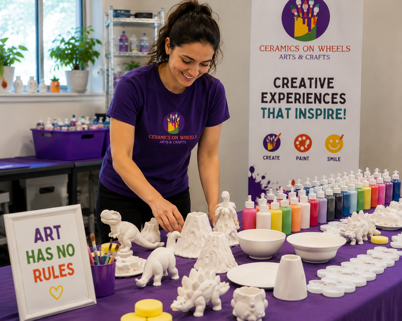 A woman smiling while arranging ceramic dinosaur sculptures and volcano models on a table at an arts and crafts event. The table is covered with a purple tablecloth, and there are numerous bottles of paint, bowls, and painted ceramic pieces. A colorful sign reads "Art Has No Rules," with a smaller poster in the background promoting ceramics arts and crafts with the slogan "Creative Experiences That Inspire!"