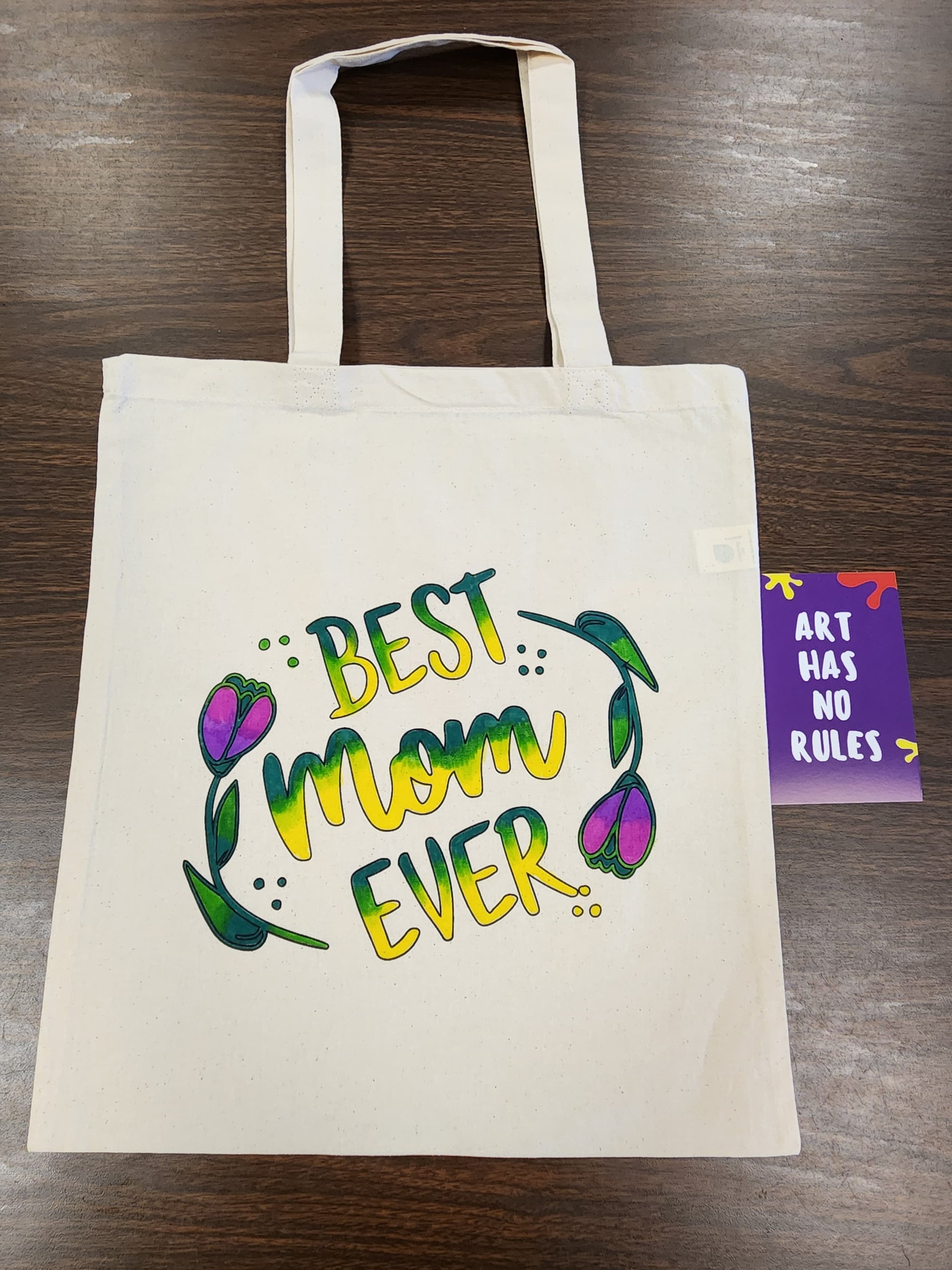 Canvas tote bag with colorful lettering that reads 'Best Mom Ever' surrounded by floral illustrations. A purple art tag saying 'Art Has No Rules' is attached to the bag.