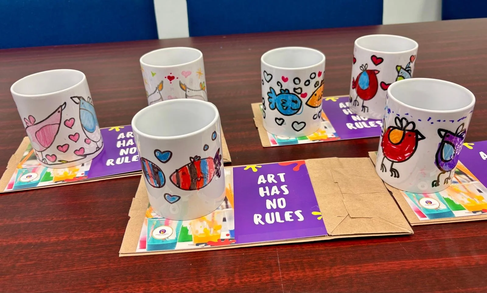 Six white mugs with colorful, cartoon-style bird and heart drawings placed on paper placemats that read "ART HAS NO RULES" in white text on purple backgrounds, on a dark wood table.