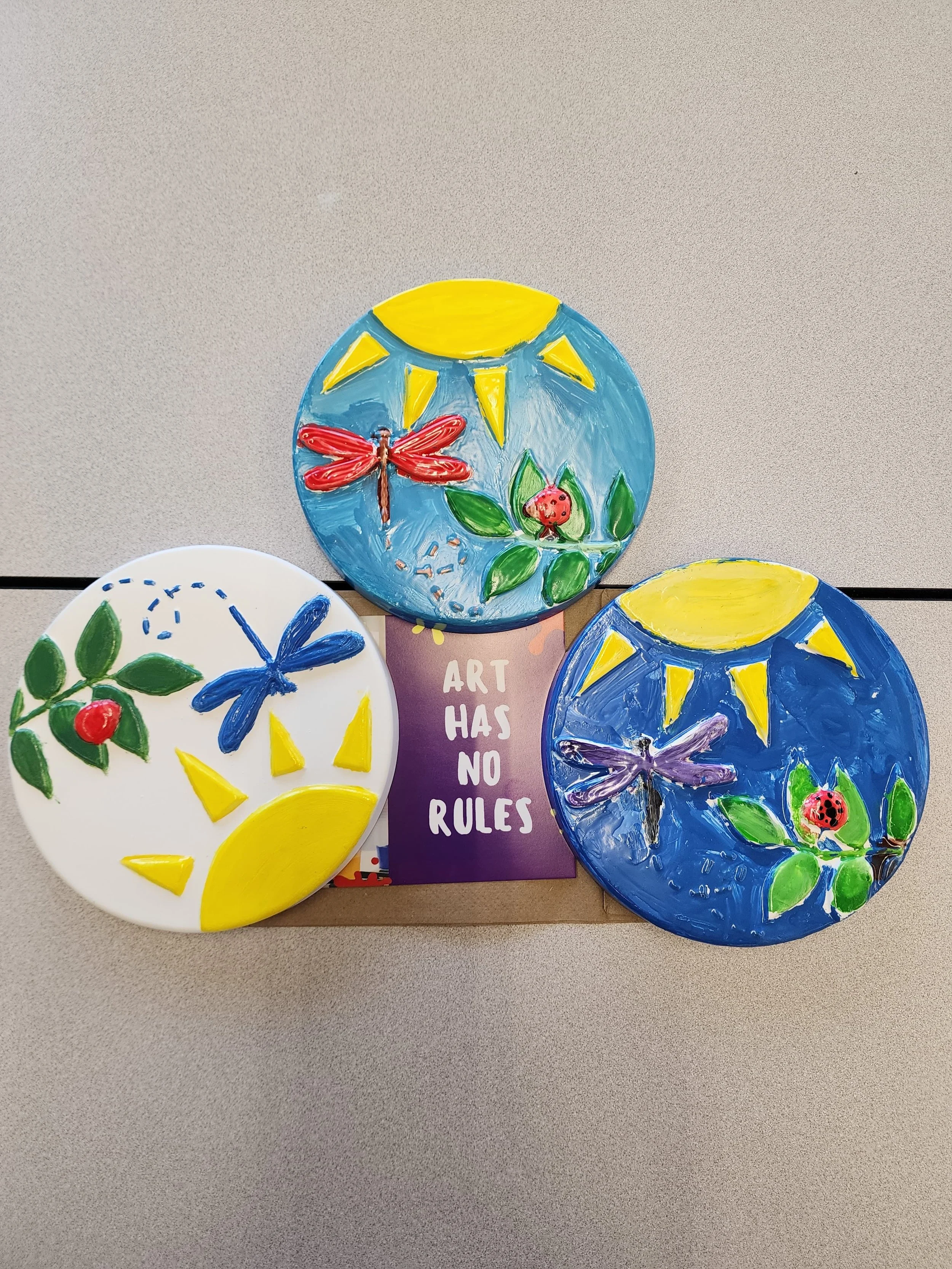 Painted decorative plates with colorful dragonfly, ladybug, and nature-themed designs on a table, with a sign that reads "Art Has No Rules."