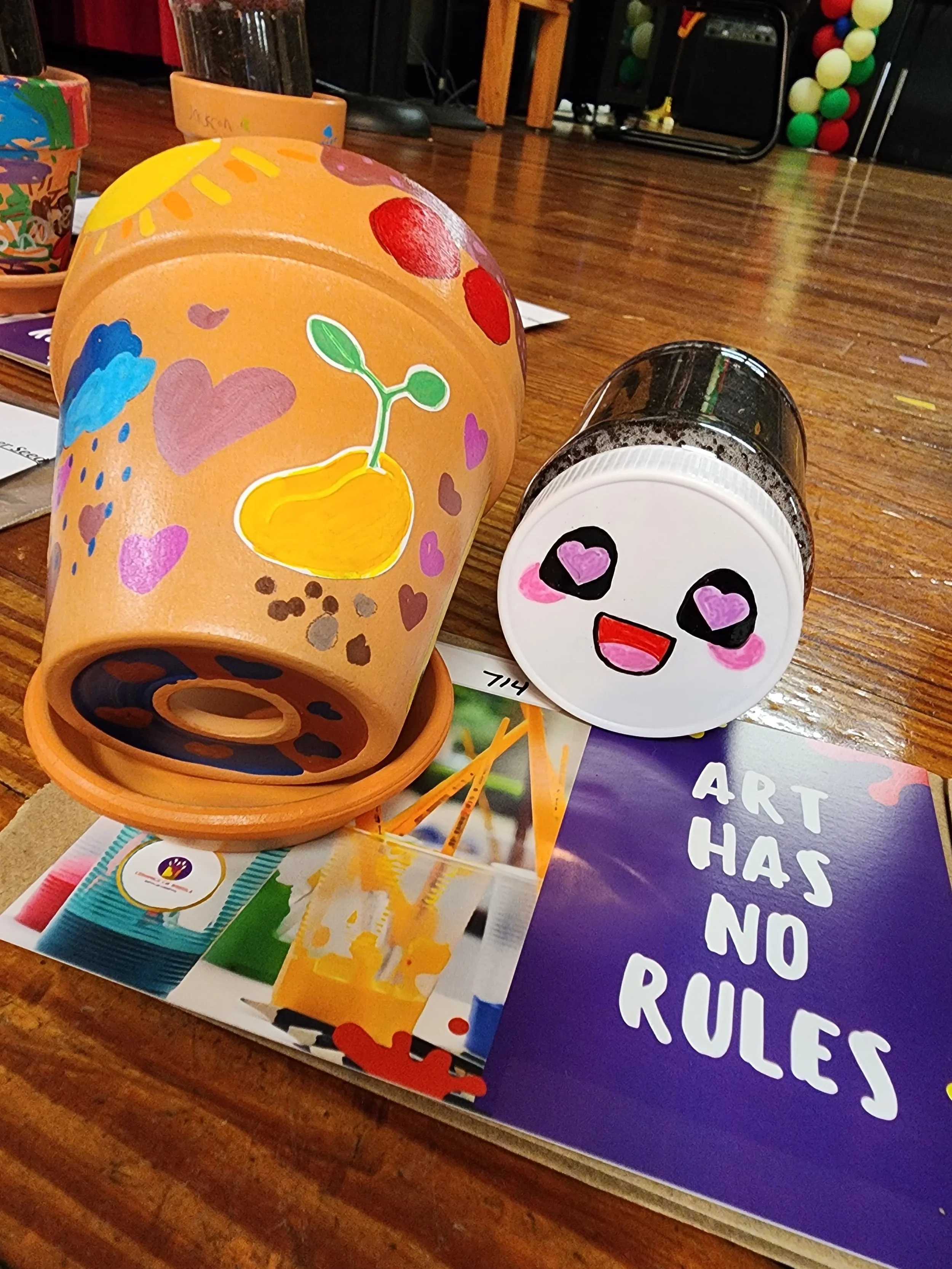 Colorful paper cups with drawn decorations and messages, including a smiley face with heart eyes and a potted plant drawing, on a wooden table with promotional materials for an art event.