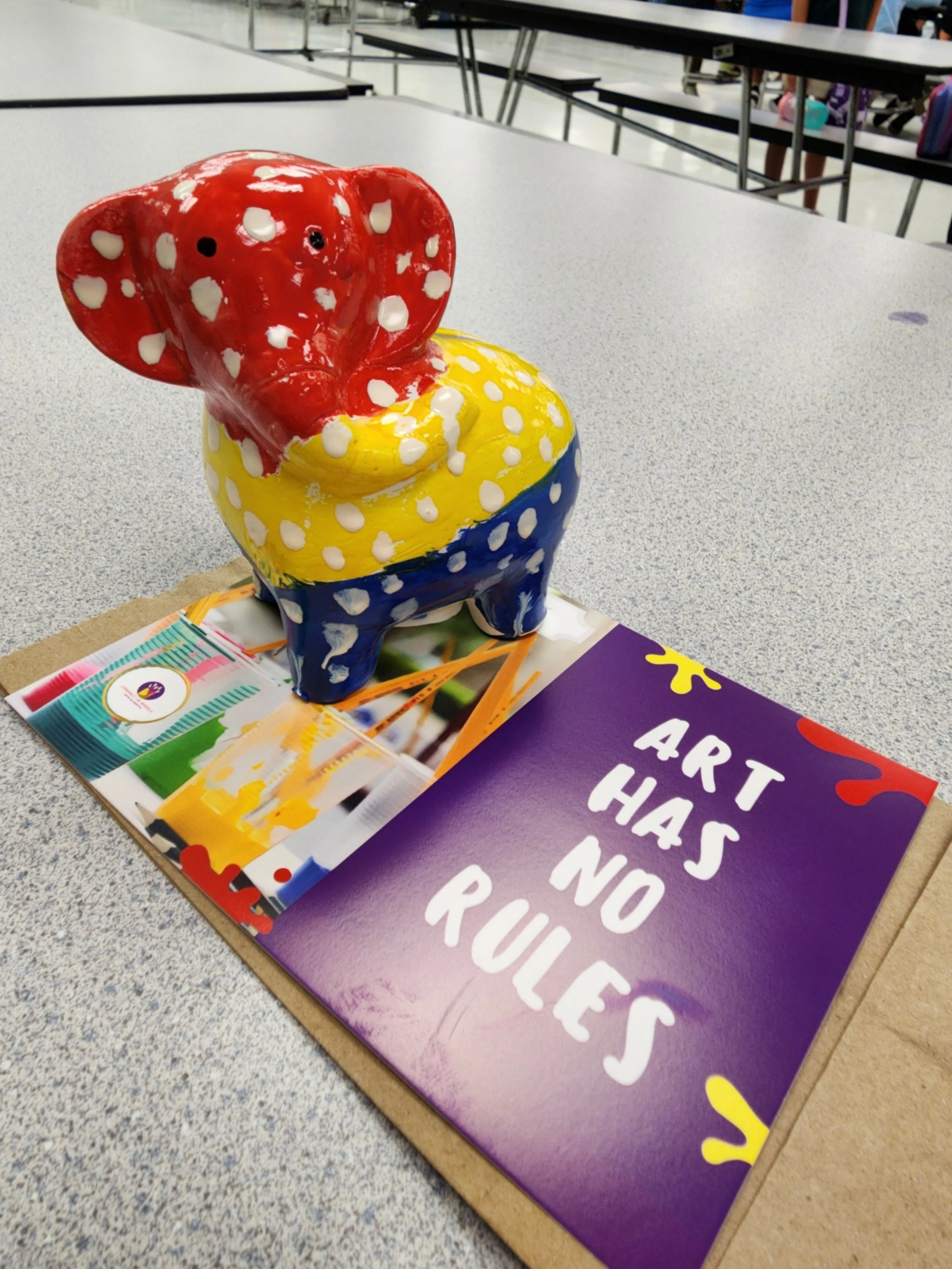 Colorful ceramic elephant sculpture with a red head, yellow middle, and blue base, placed on a table with a colorful art booklet and a purple sign that reads 'ART HAS NO RULES'.