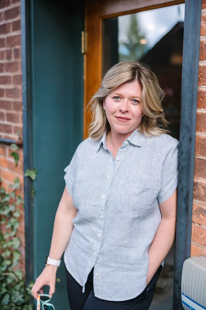 A woman with blonde hair stands casually against a brick wall, wearing a light blue button-up shirt. She holds sunglasses, exuding a relaxed and friendly vibe.