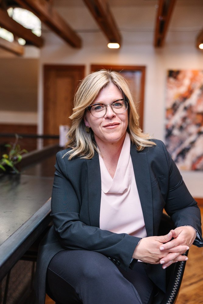 Katharine a professional coach with blonde hair and glasses sits confidently in an office. She wears a dark blazer and light blouse, exuding a calm focus.
