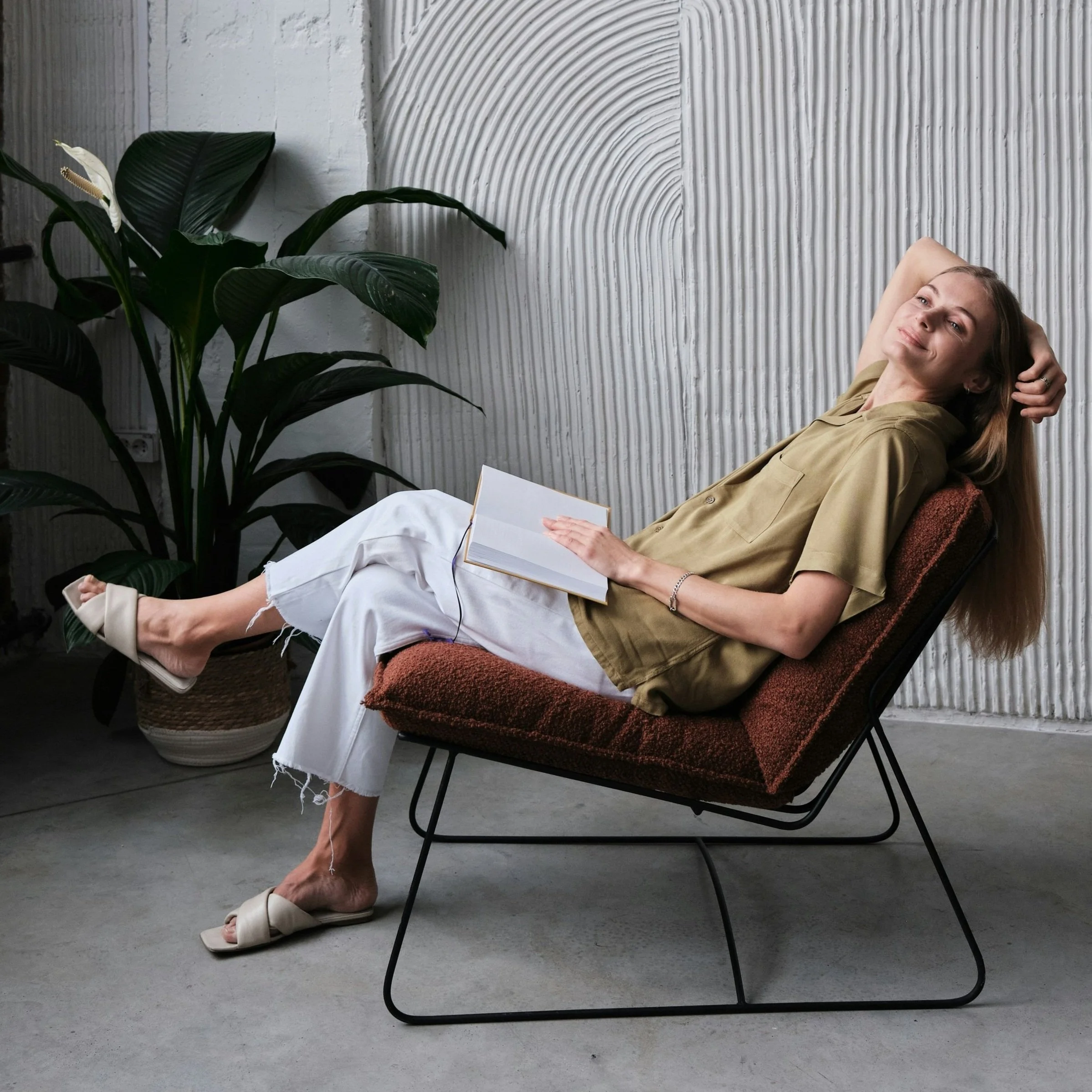 A person relaxing in a chair with a book, surrounded by a modern textured wall and a potted plant.