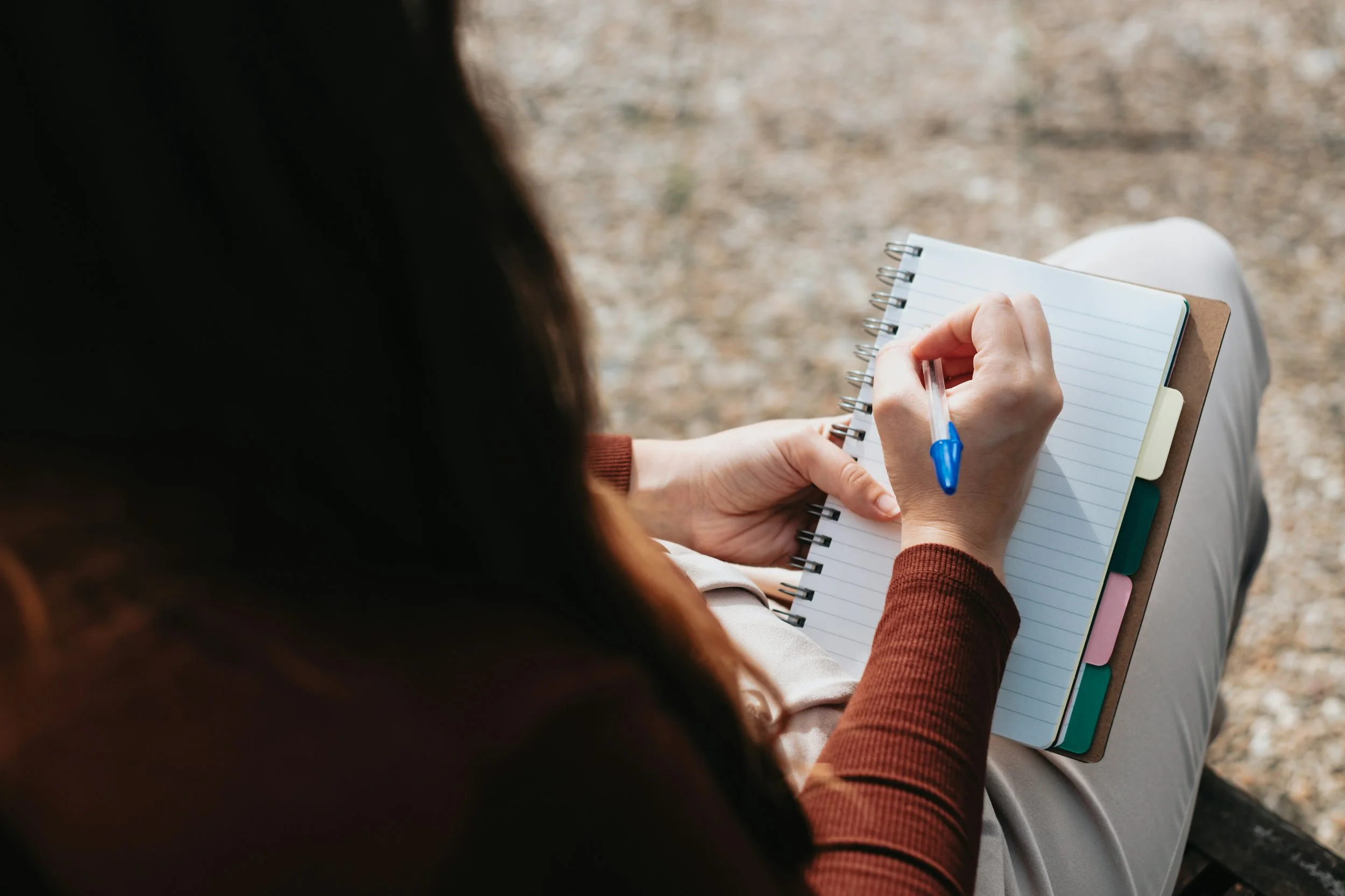 Person writing in a spiral notebook with a blue pen, sitting on a bench outdoors.