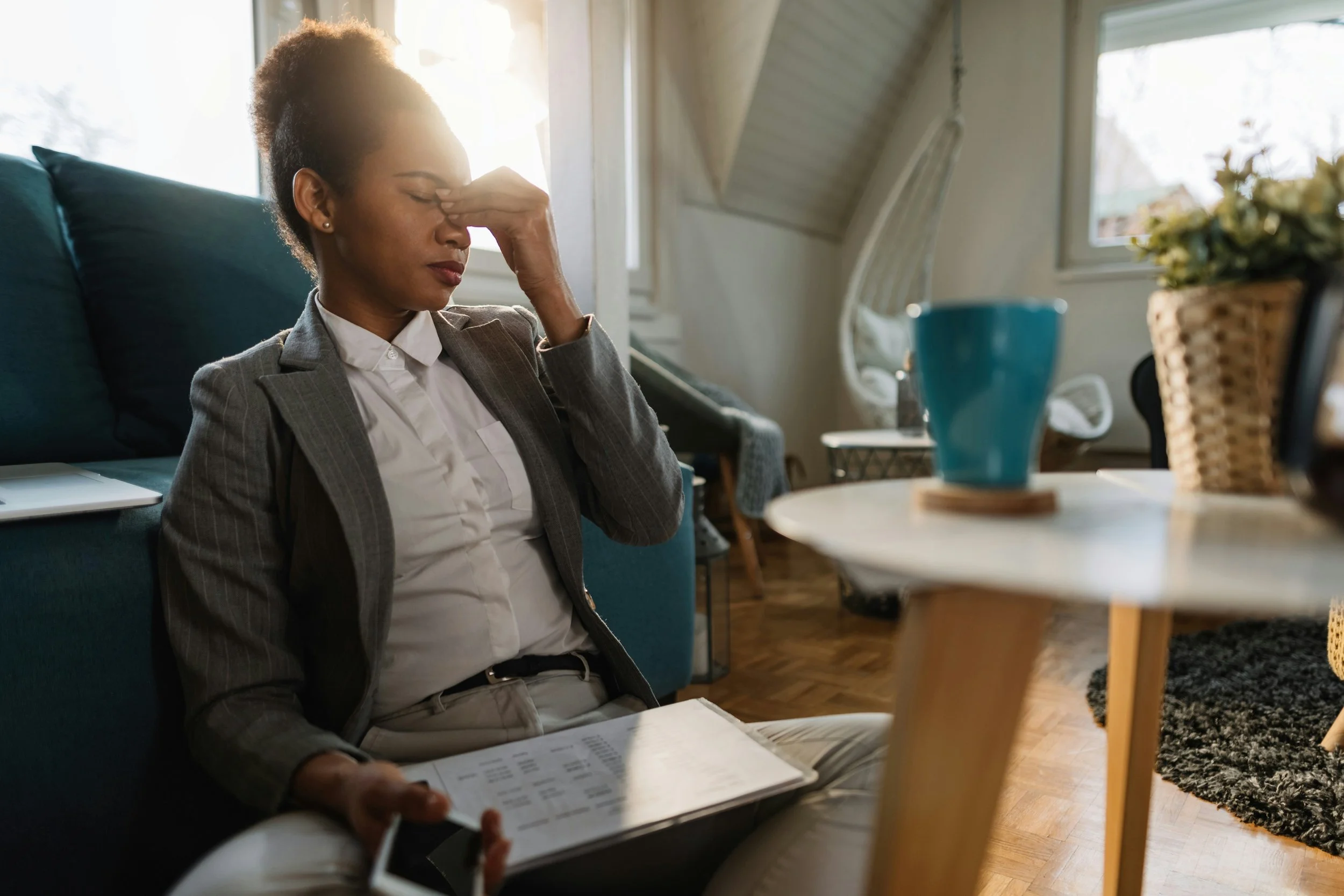 Woman in a suit sitting on the floor, pinching her nose bridge in a stressed manner, with a notebook and smartphone in hand, in a home setting.