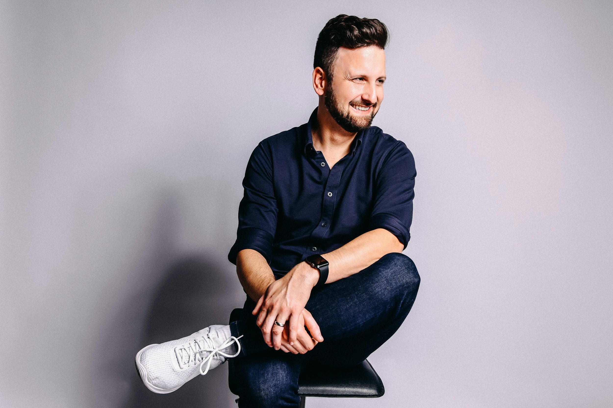 Man sitting on a stool, wearing a navy blue shirt, dark jeans, and white sneakers against a gray background.