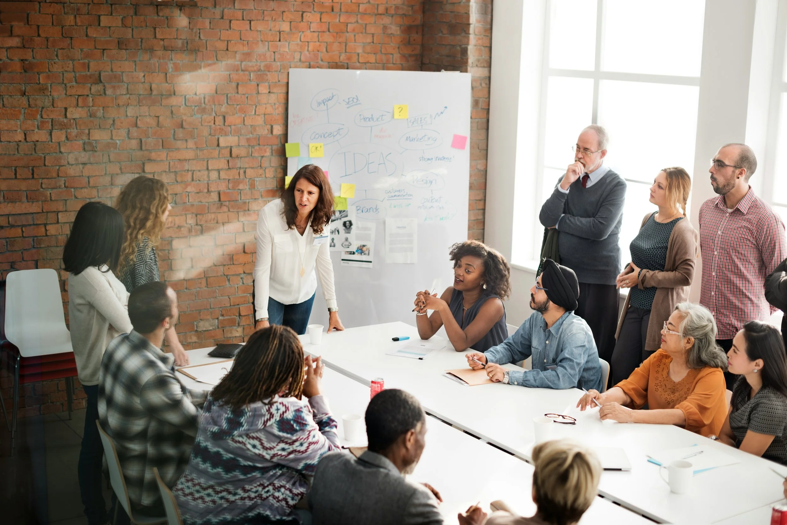 A diverse group of people in a business meeting around a conference table with a whiteboard covered in notes. A woman in a white shirt is leading the discussion. The room has a brick wall and large windows.