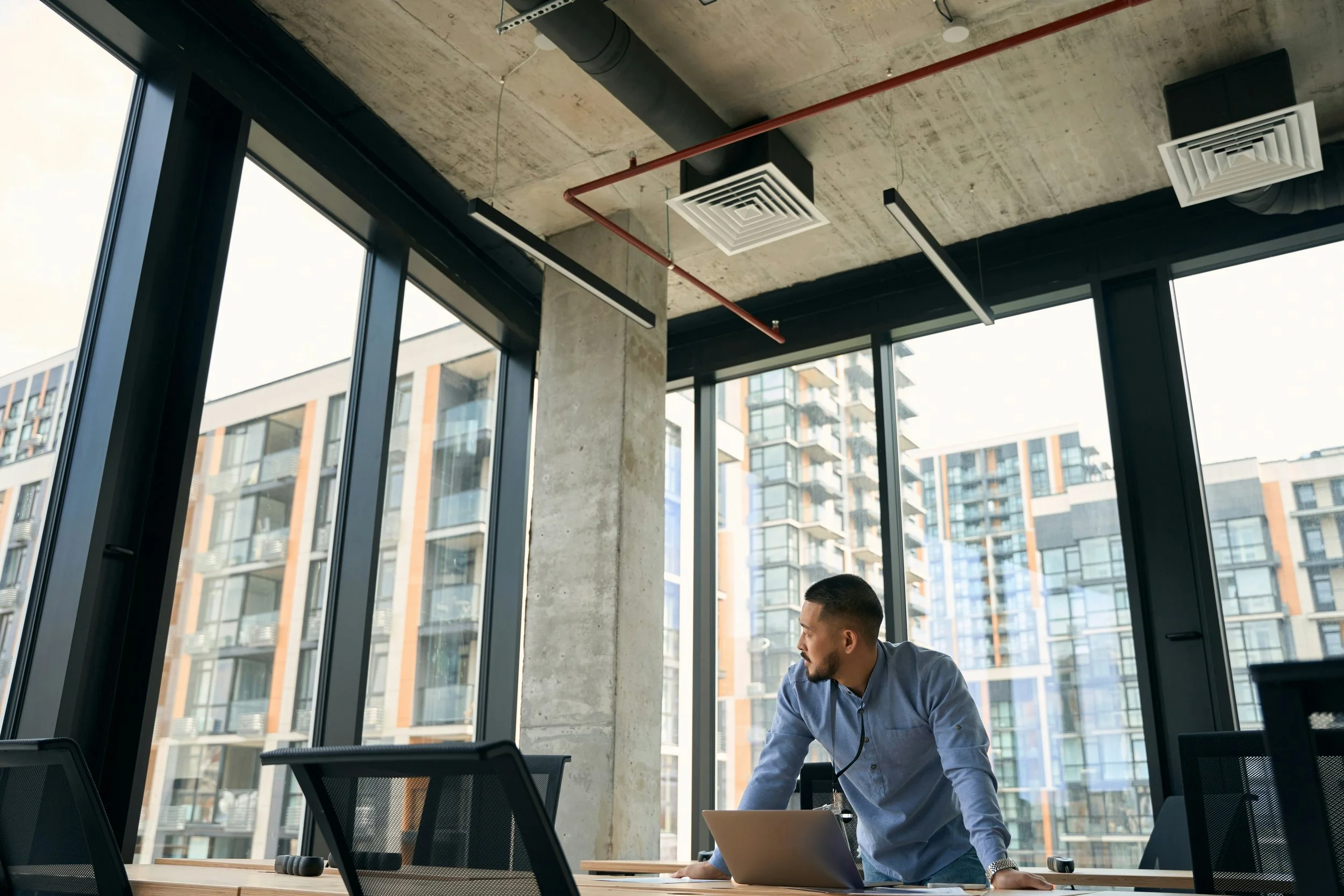 Man in a modern office with large windows looking out at a cityscape with high-rise buildings.