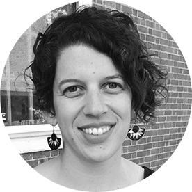 Black and white portrait of a person with short curly hair, wearing earrings, smiling, with a brick wall background.
