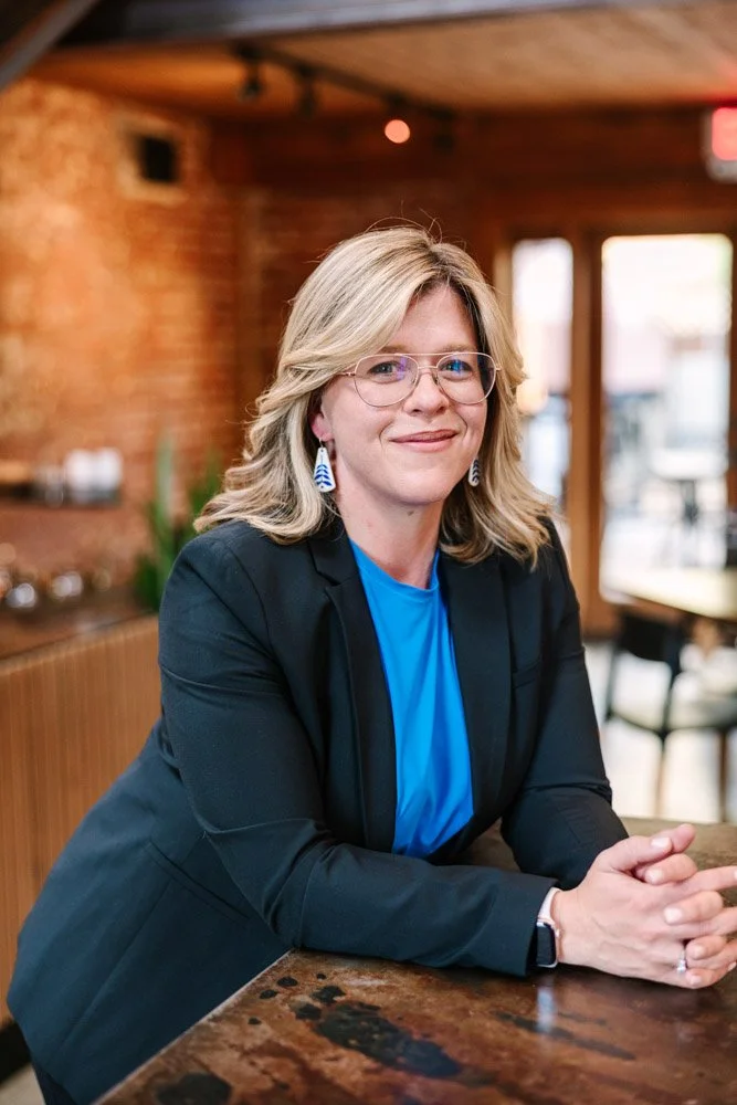 Katharine a blonde woman in glasses and a blazer leans on a wooden table, smiling confidently. Brick-walled café setting with soft lighting conveys a warm tone.