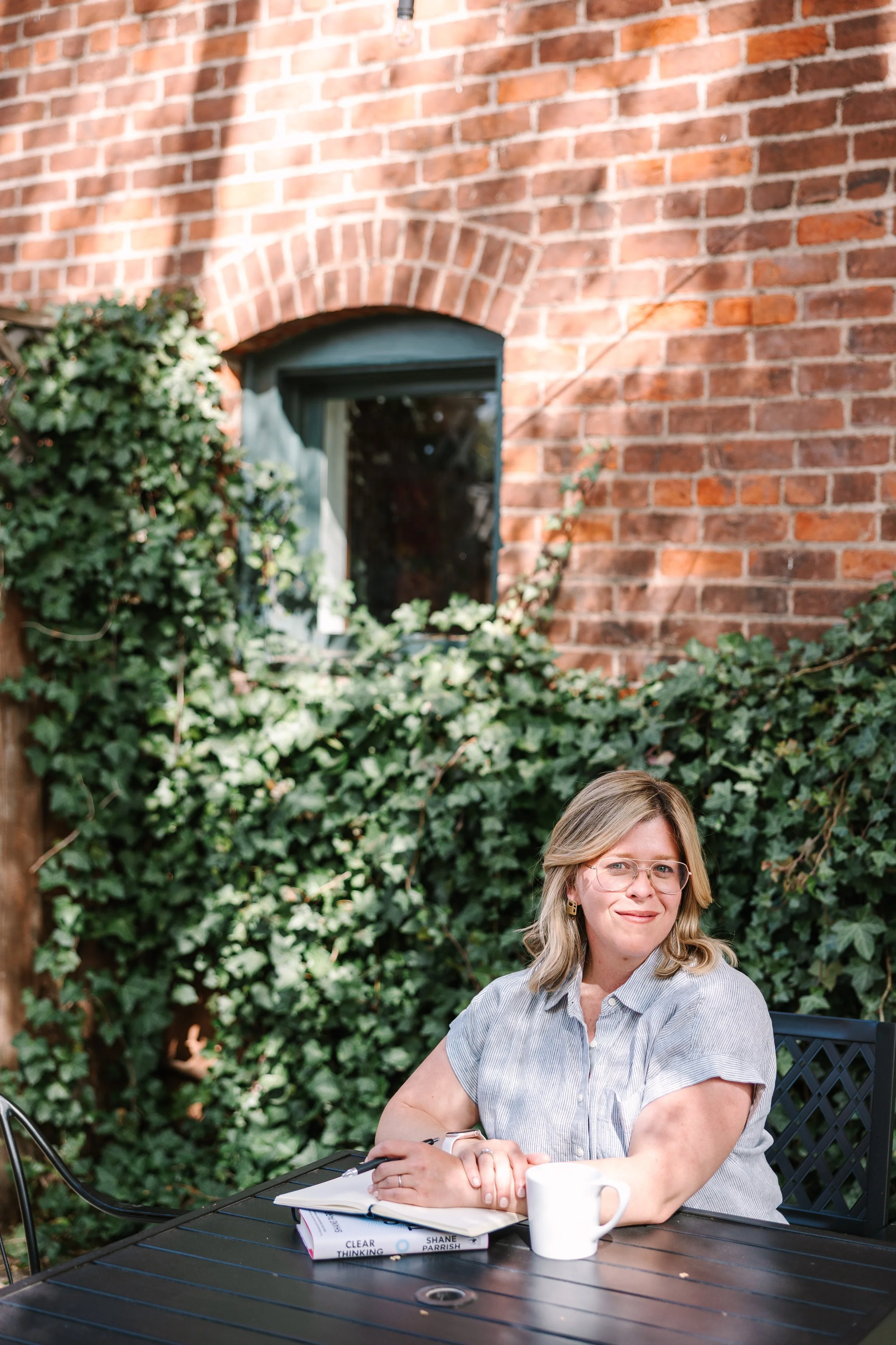 Katharine, a blonde woman sitting at a patio table against a brick wall covered in ivy, holding a coffee mug. Sunlight creates a peaceful, warm atmosphere.