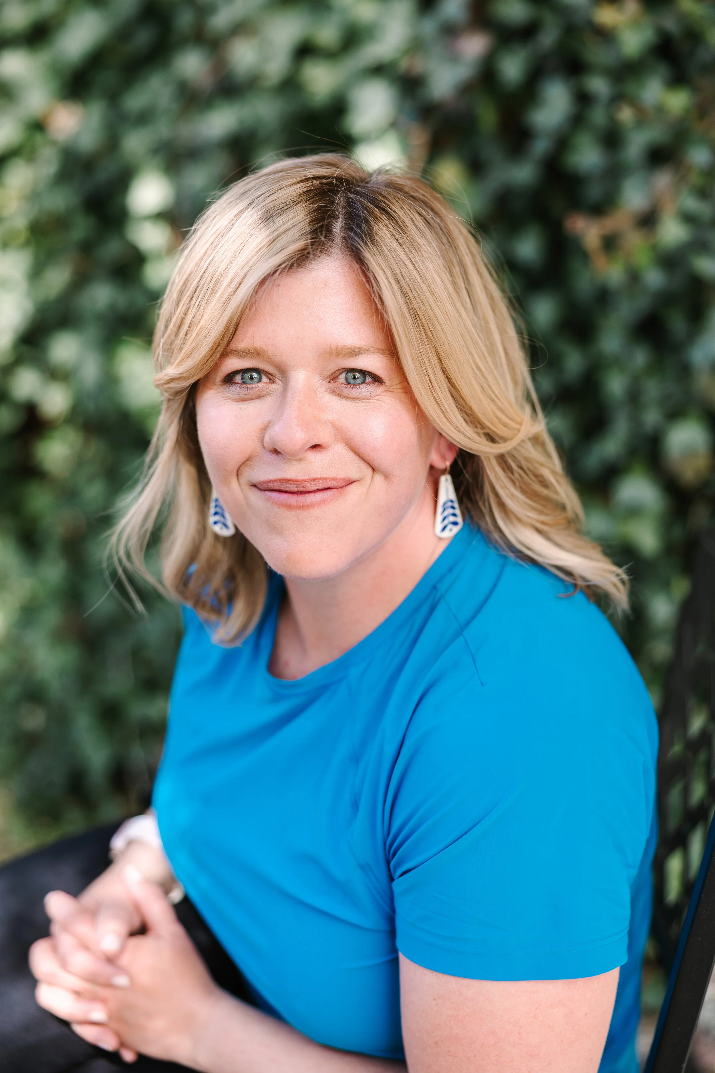 A woman with blonde hair wearing a blue shirt, sitting outdoors with a blurred green leafy background, smiling and looking at the camera.