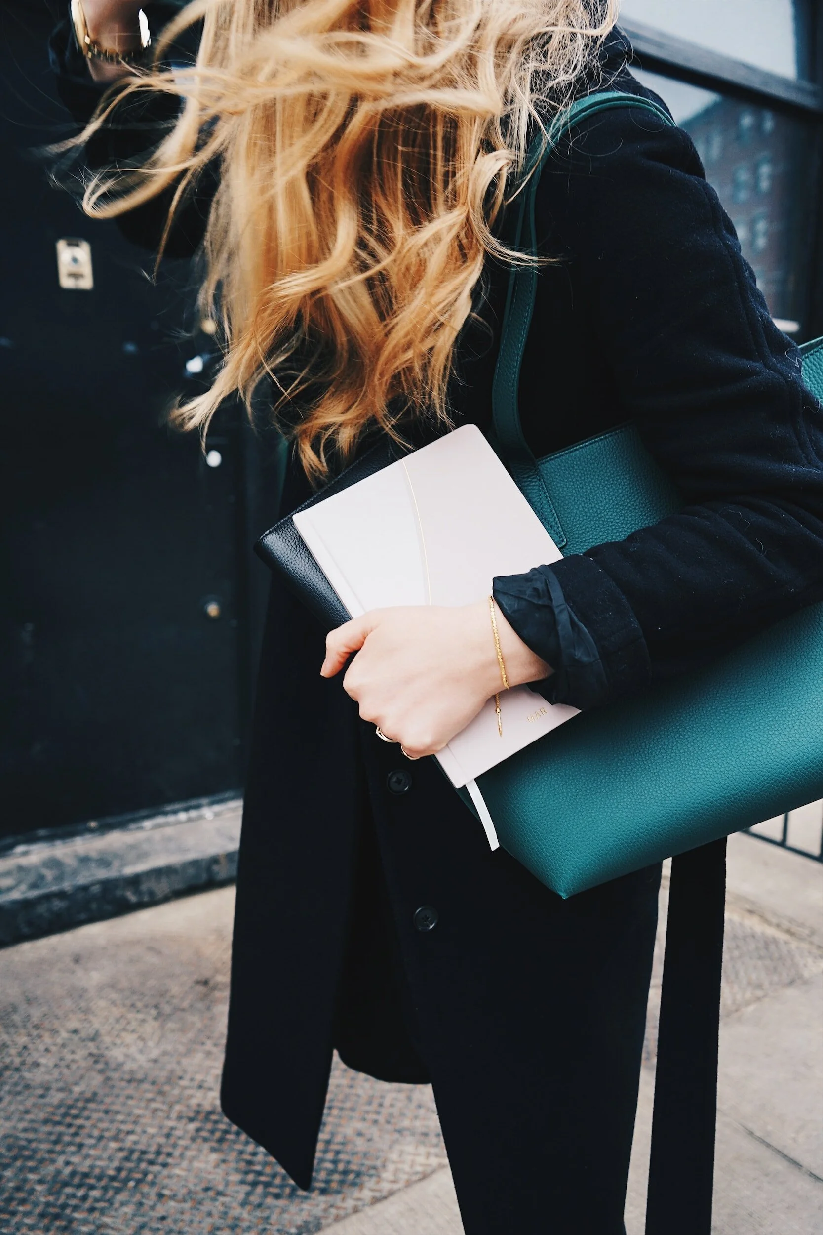 Woman with blonde hair holding a notebook and a teal handbag, wearing a black coat outdoors.