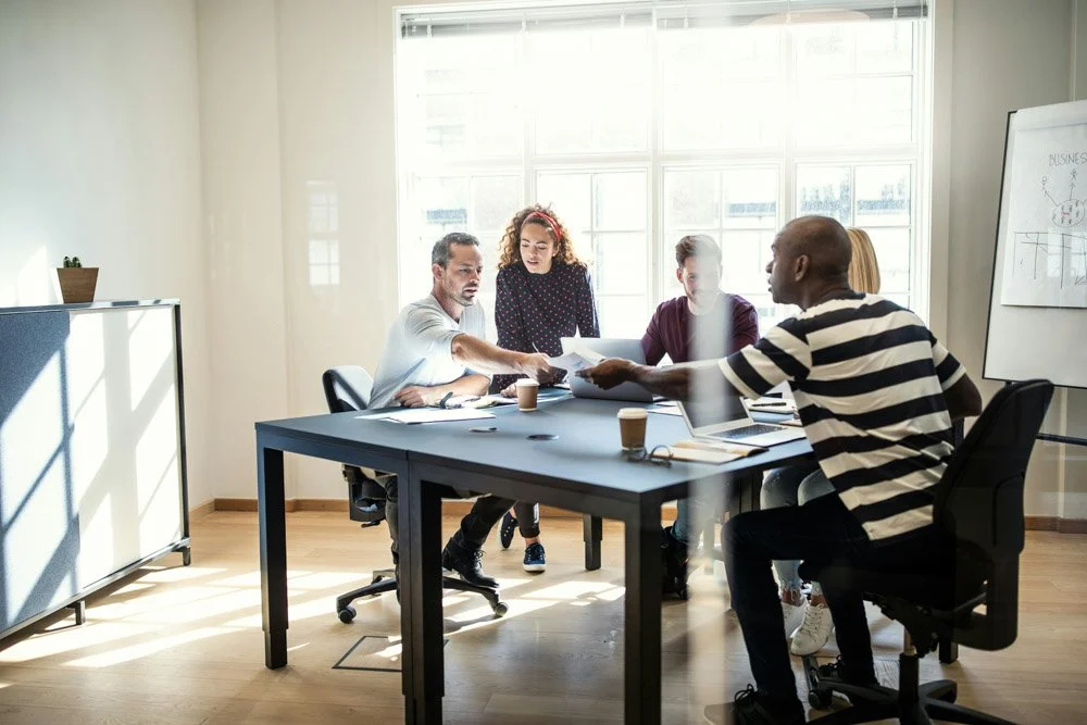 A diverse group of four professionals collaborates in a bright office, gathered around a table with papers and coffee cups, focused on solving a problem..