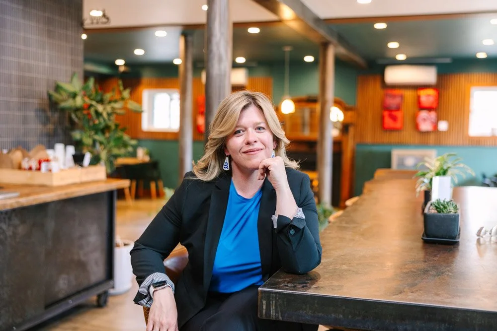 Katharine a woman with blonde hair, wearing a blue shirt and black blazer, sits confidently at a wooden table in a warmly lit, modern office space.
