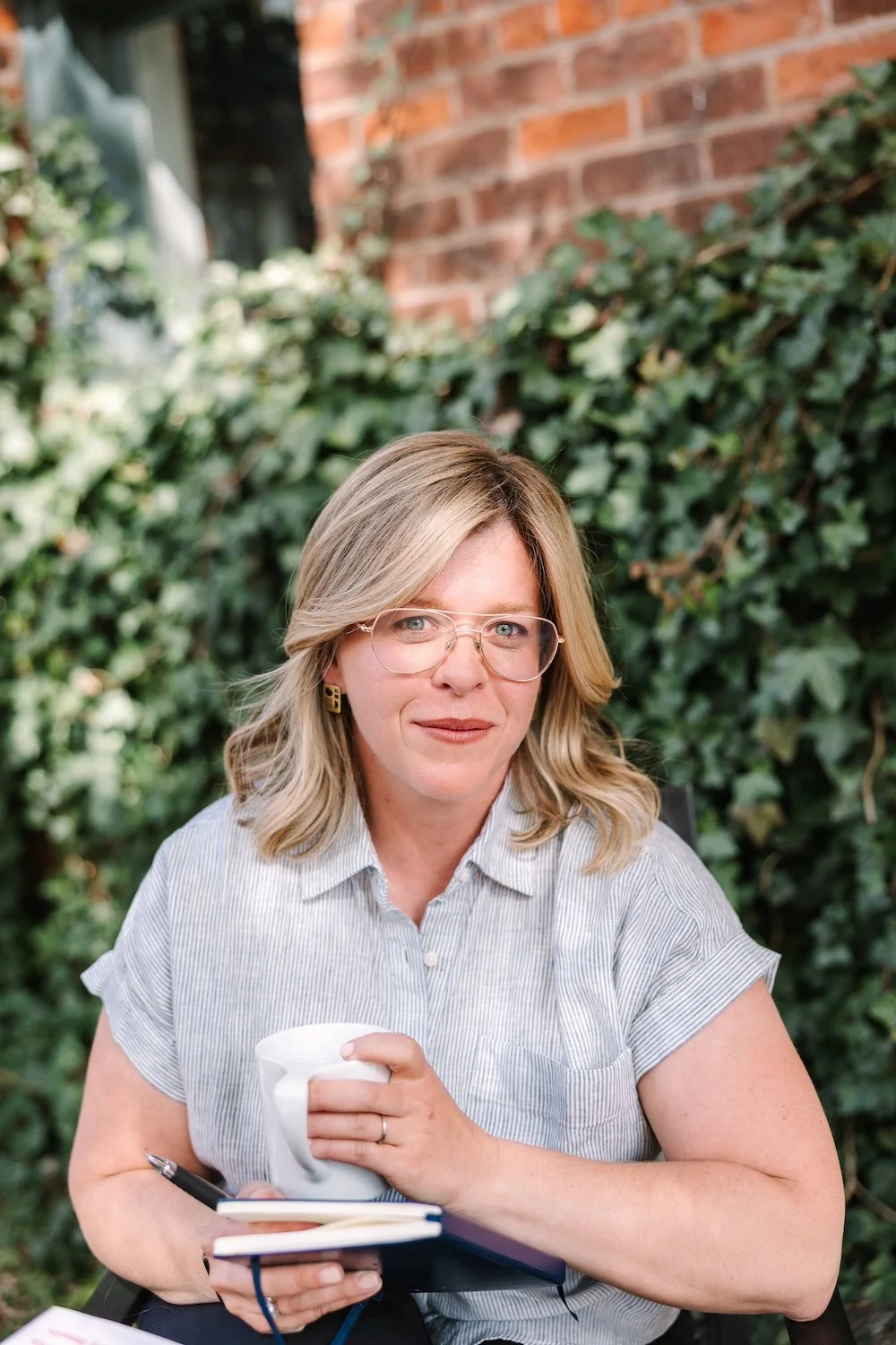 Woman with blonde hair and glasses, smiling while holding a coffee cup and notebook, sits outdoors against a brick wall covered in green ivy.