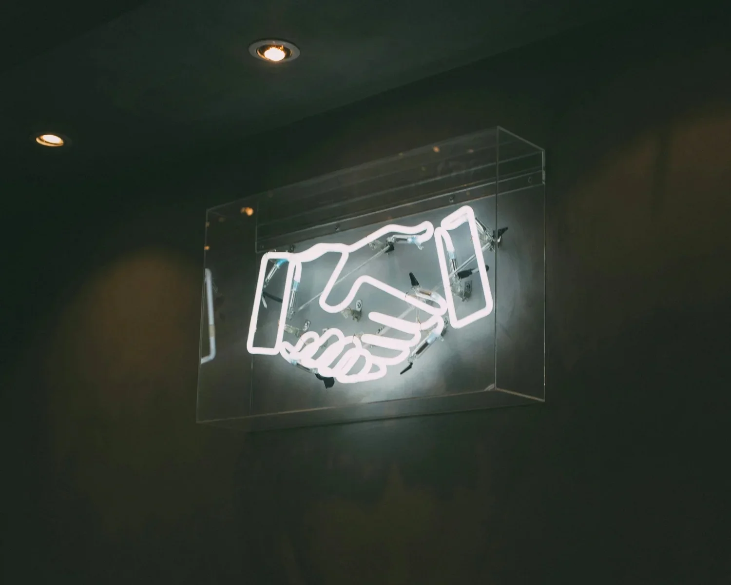 Neon sign of two hands shaking, glowing brightly against a dark wall. The image conveys a theme of partnership and agreement.
