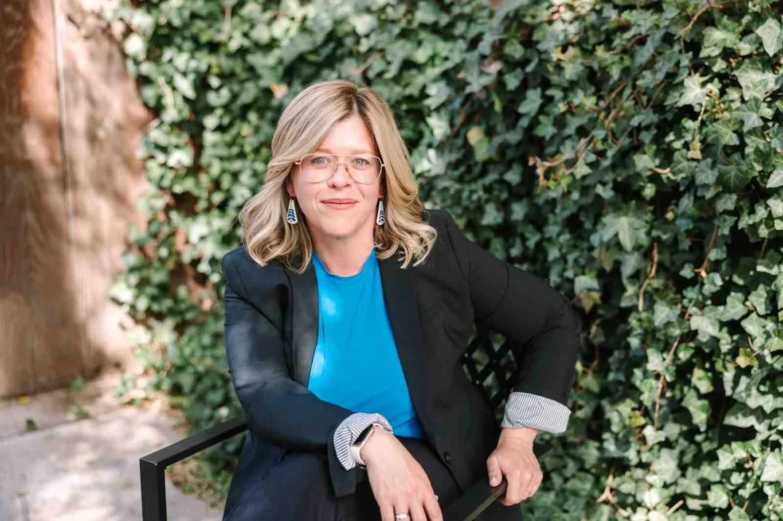 Katharine a woman with glasses and blonde hair sits confidently outdoors against a lush ivy backdrop, wearing a black blazer and blue top, exuding professionalism.