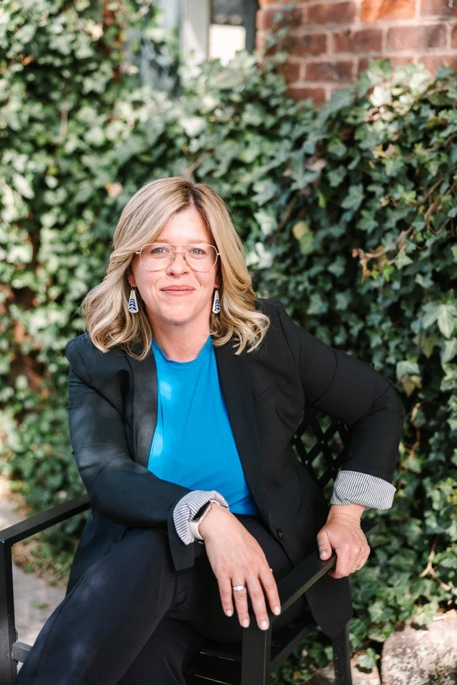 Katharine a woman with blonde hair and glasses sits confidently on a black chair. She's in a black blazer and blue shirt, with a background of lush green ivy.