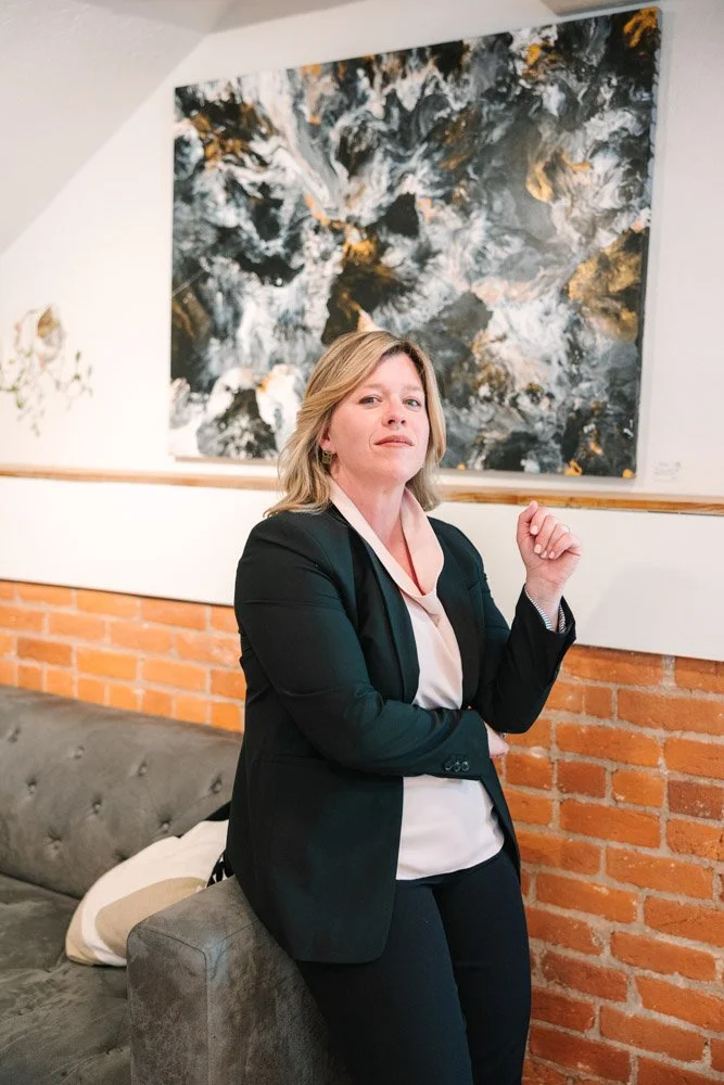 A woman in a black suit leans against a gray couch, exuding confidence. Behind her is a dynamic abstract painting with dark swirls and hints of gold.