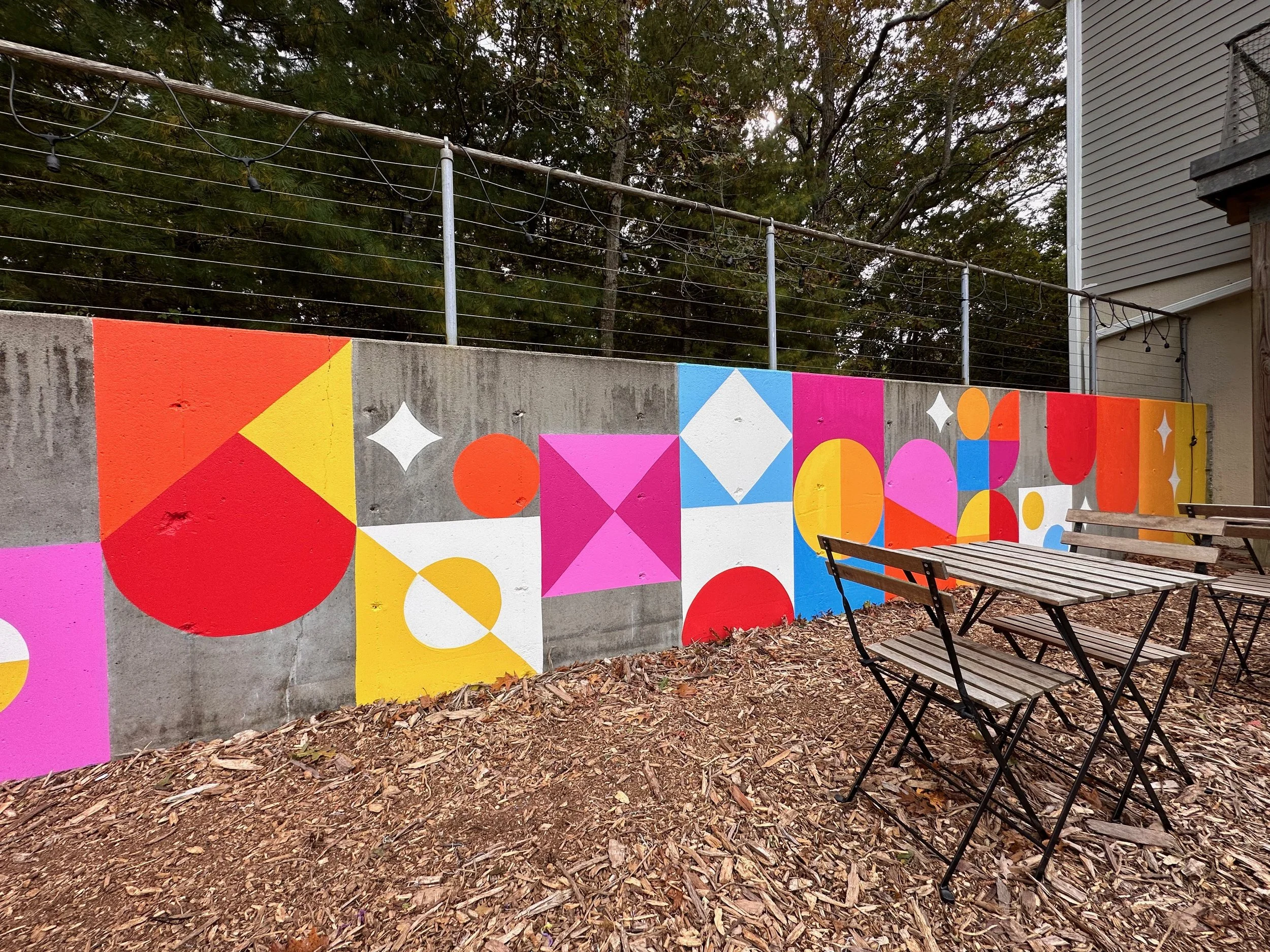 SHINE Studio Retaining Wall: a bright, geometric installation transformed this concrete retaining wall to a cheerful backdrop to the outdoor seating and playground areas.  One of a number of mural installations commissioned by the Studio of local art