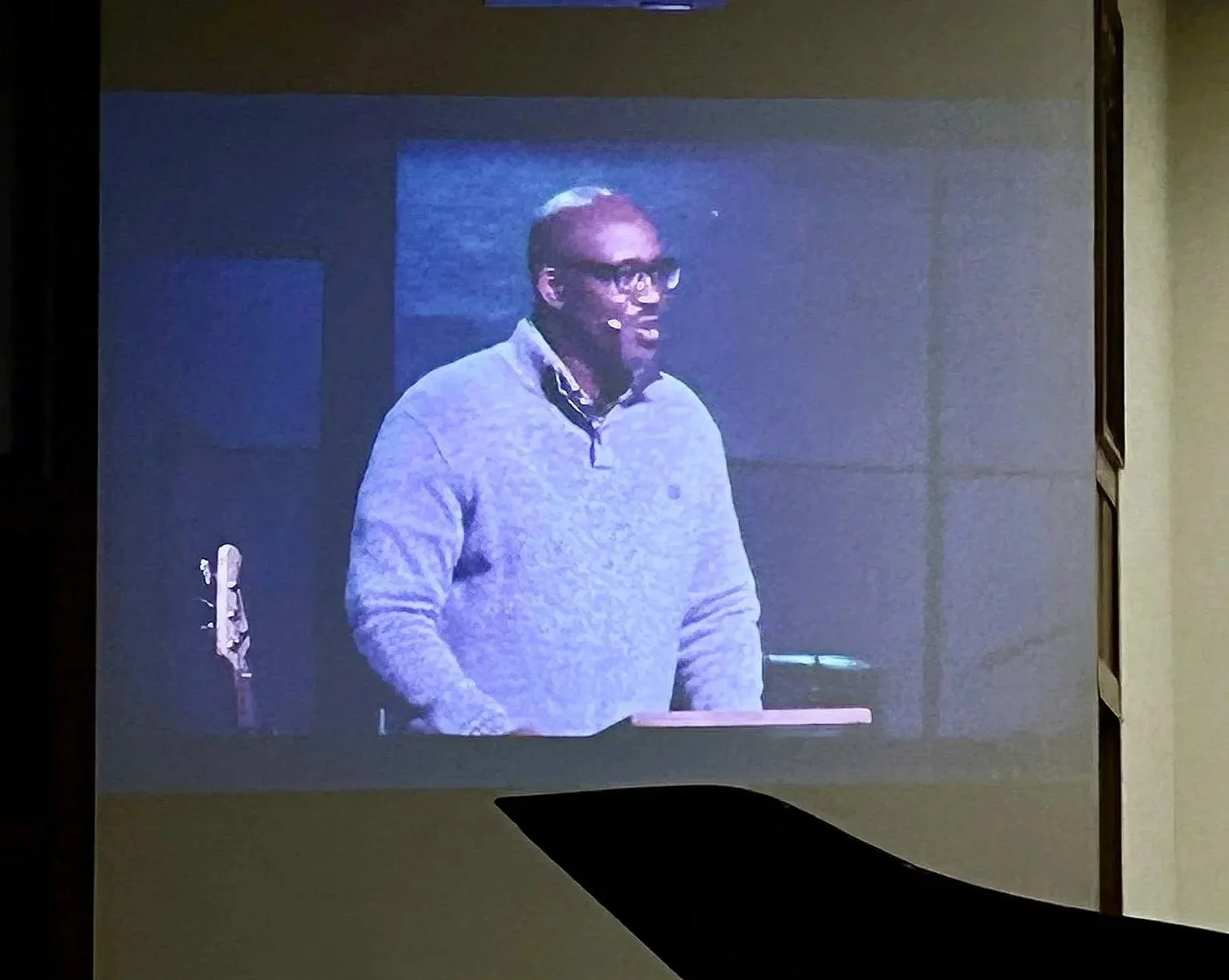 A man with glasses speaking on a stage or platform, with a guitar in the background.