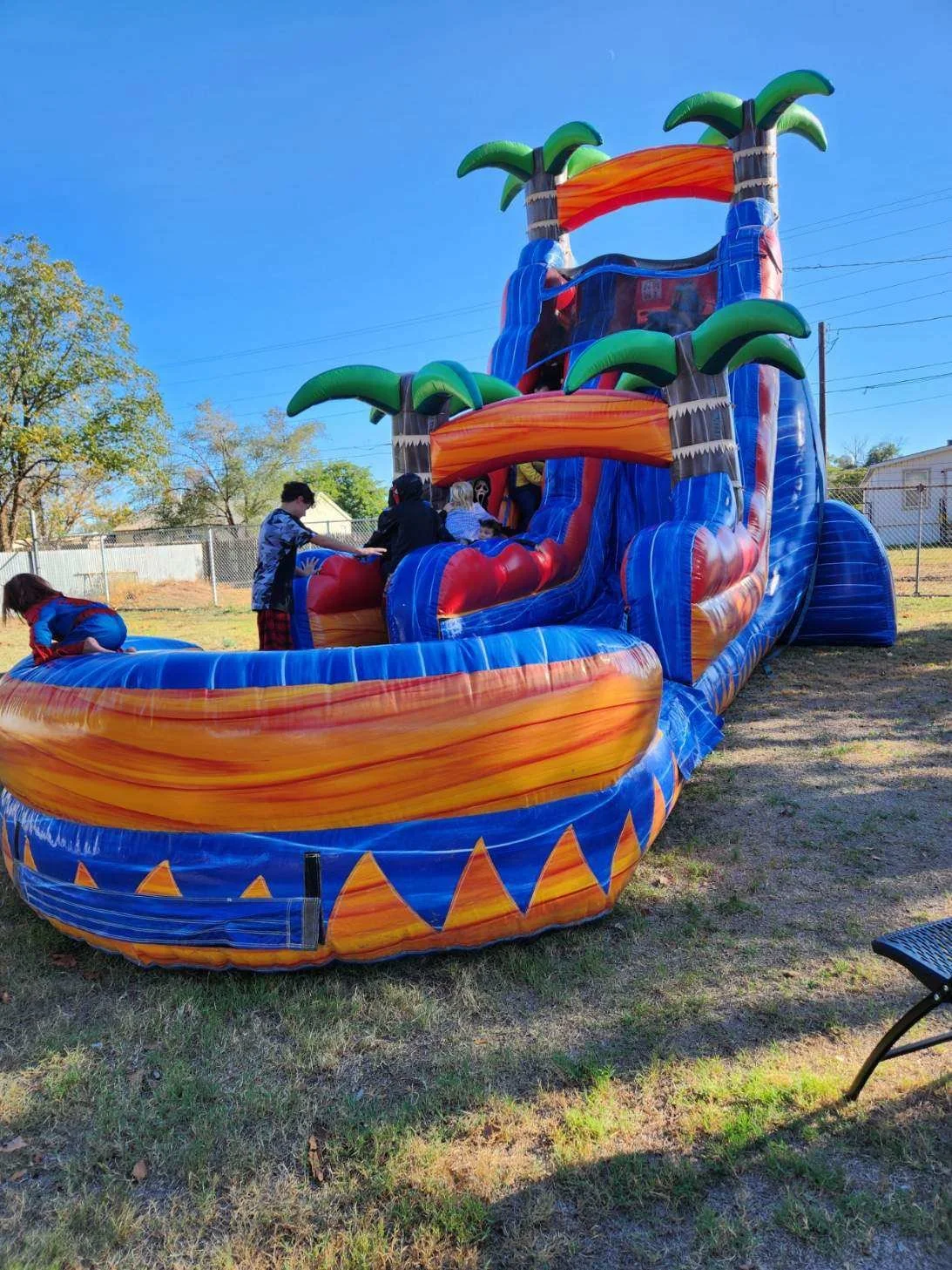 Inflatable water slide designed to look like a tropical island with palm trees, water cascading down the slides, on a grassy outdoor area with children playing.