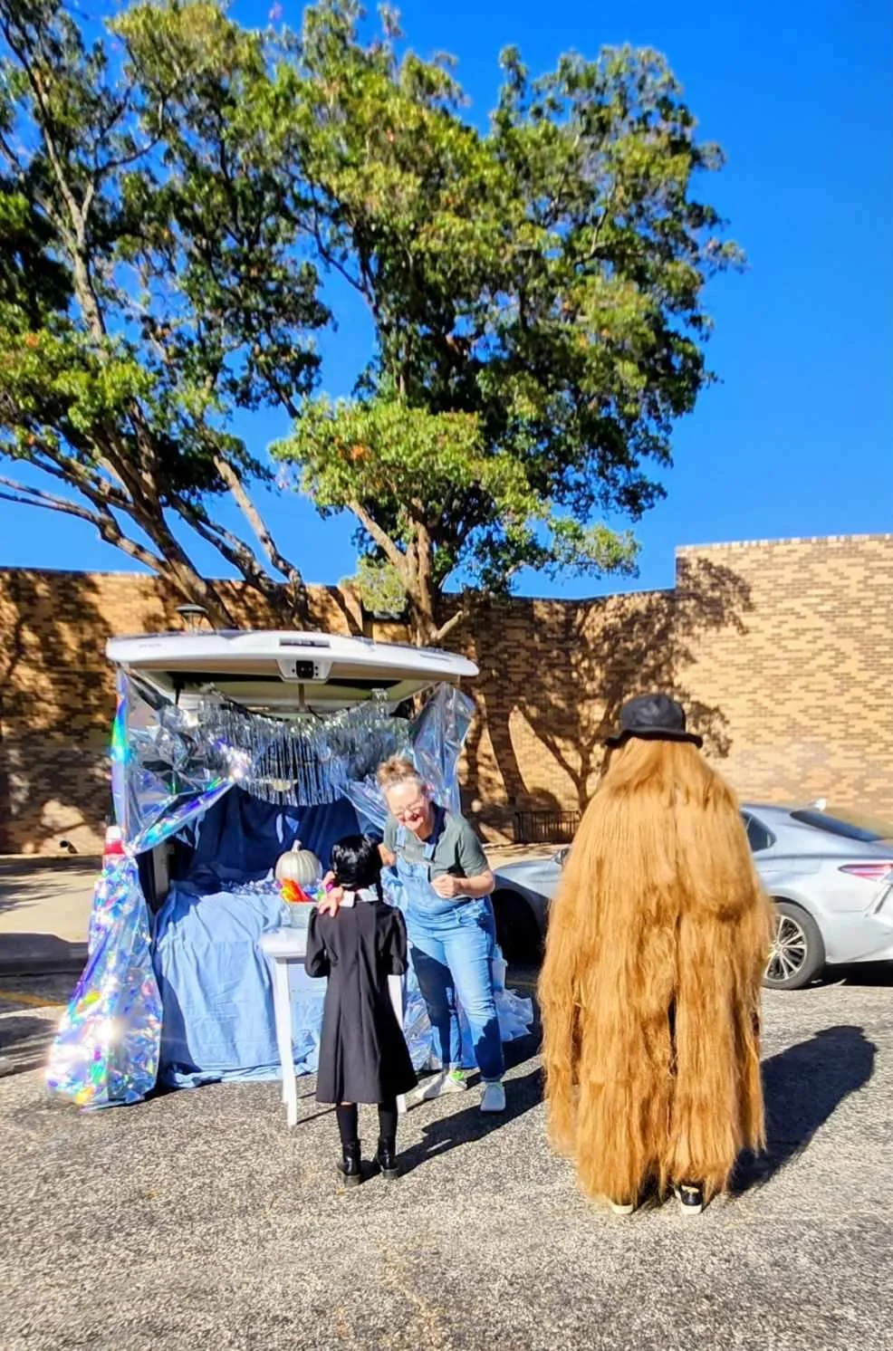 A Halloween scene with a woman and a child in a witch costume at a decorated Halloween booth, with a person dressed as a Bigfoot standing nearby, in a parking lot under a clear blue sky.