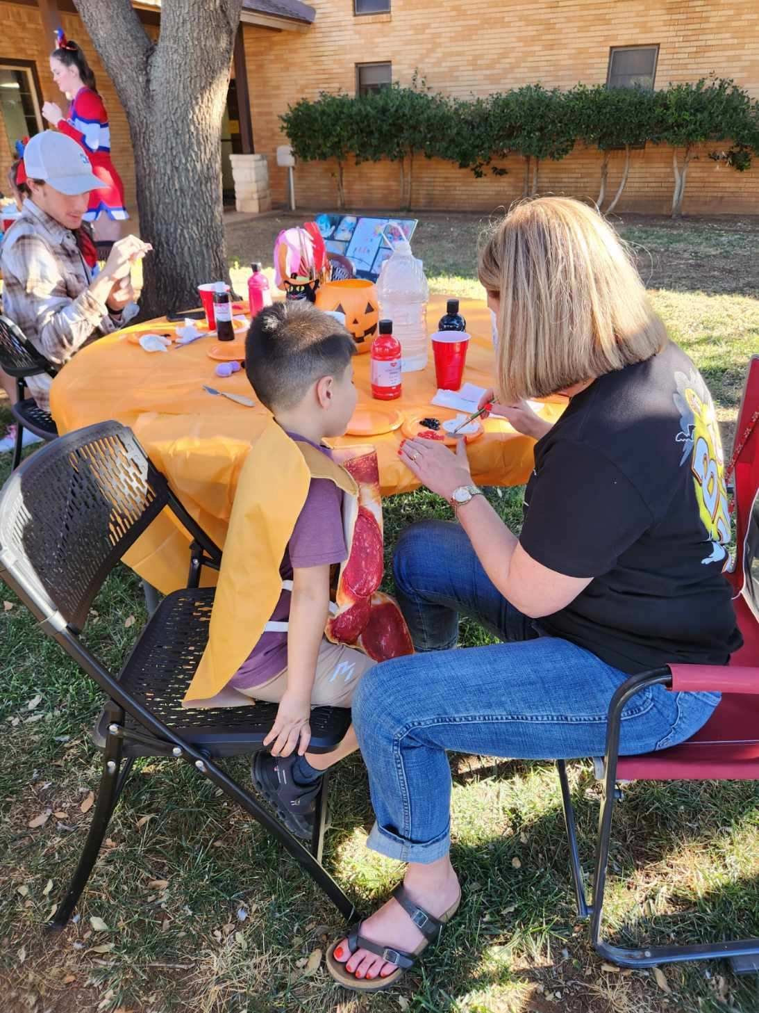 People decorating for Halloween at an outdoor party. A woman is sitting at a table with two children, applying face paint or decorations to a child. The table is covered with orange tablecloth, Halloween-themed decorations, and supplies. In the backg