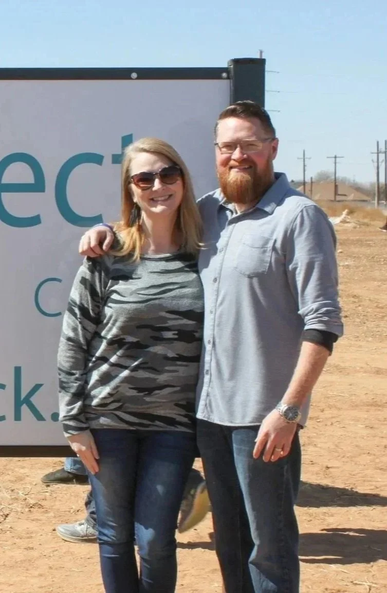 A man and woman smiling and embracing outdoors, with a large sign and power lines visible in the background.