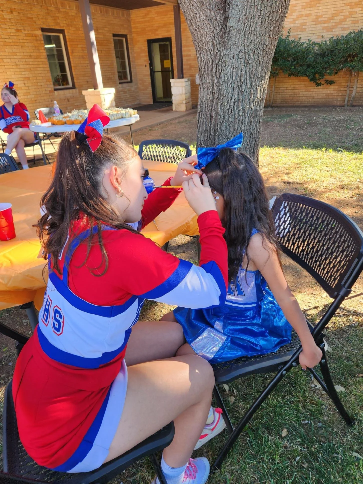 A young woman dressed in a cheerleading outfit painted stars on a young girl's face at an outdoor party under a tree, with other children and a table of snacks in the background.