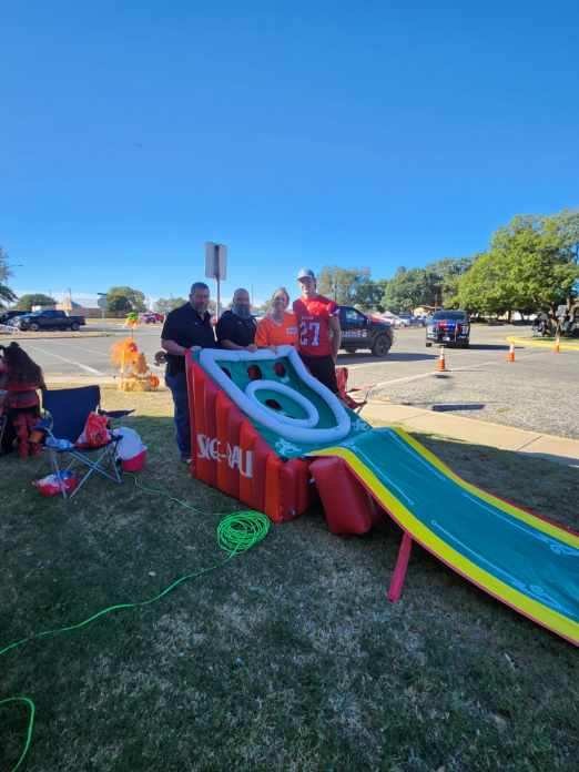 People standing next to a large inflatable slide set up on a grassy area near a parking lot with cars and trees, under a clear blue sky.