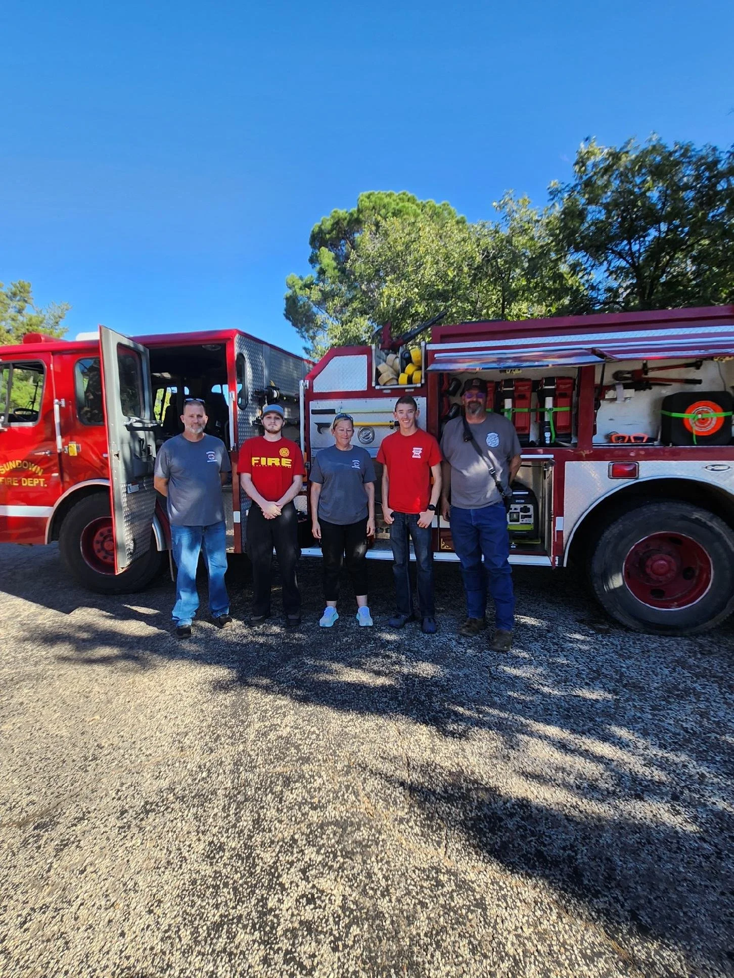 Five people standing in front of a red fire truck outdoors on a sunny day.