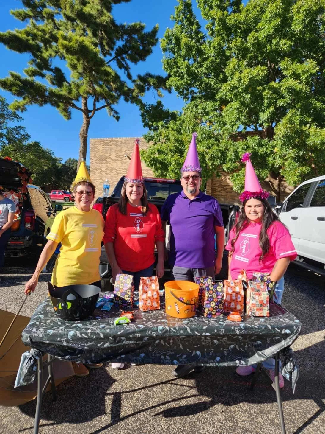 Four people dressed in colorful shirts and birthday hats standing behind a table with gift bags and Halloween-themed decorations on it, outdoors on a sunny day.