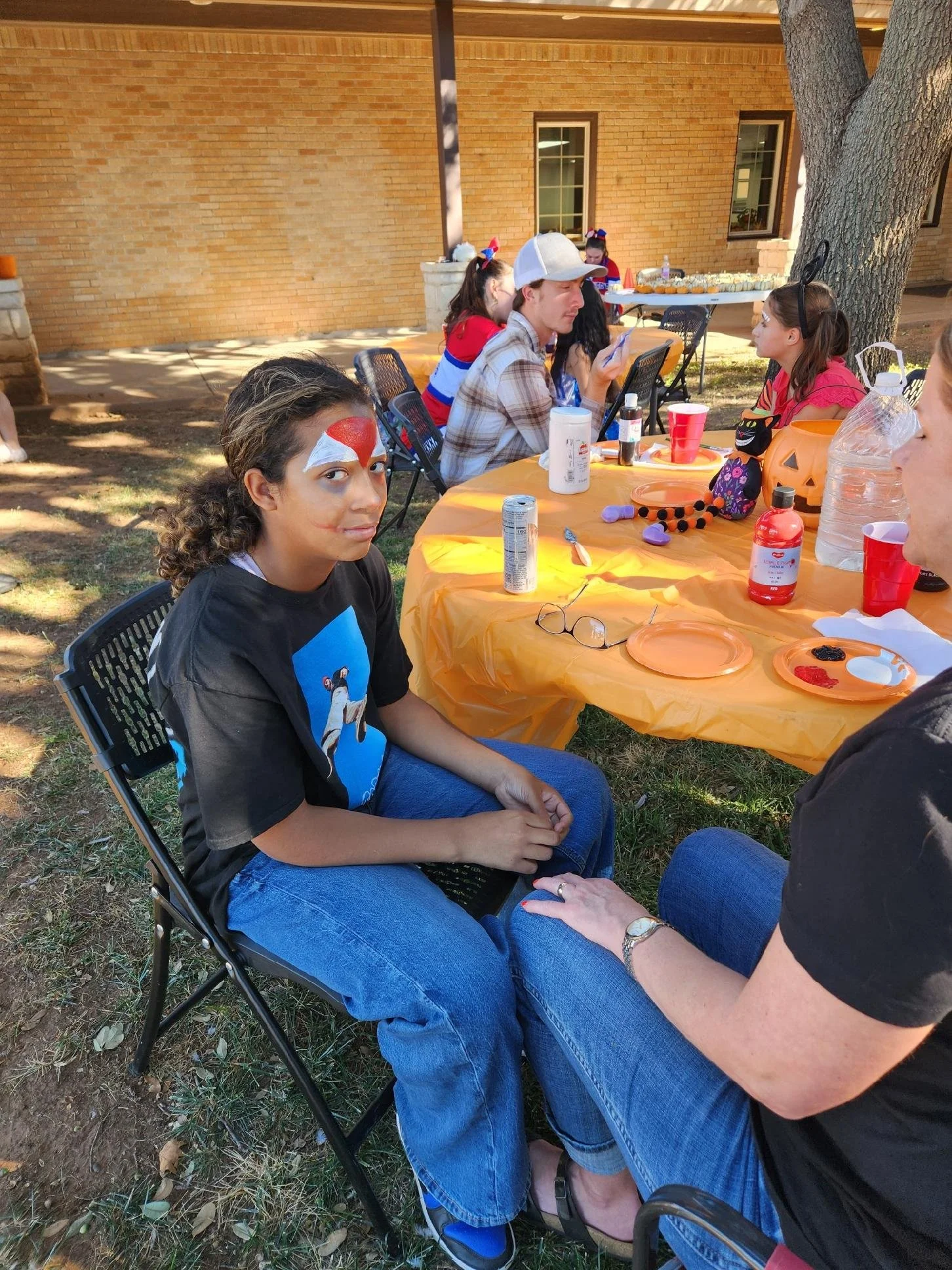 Children and adults at a Halloween-themed outdoor party, with face paint, costumes, and festive decorations on a table.