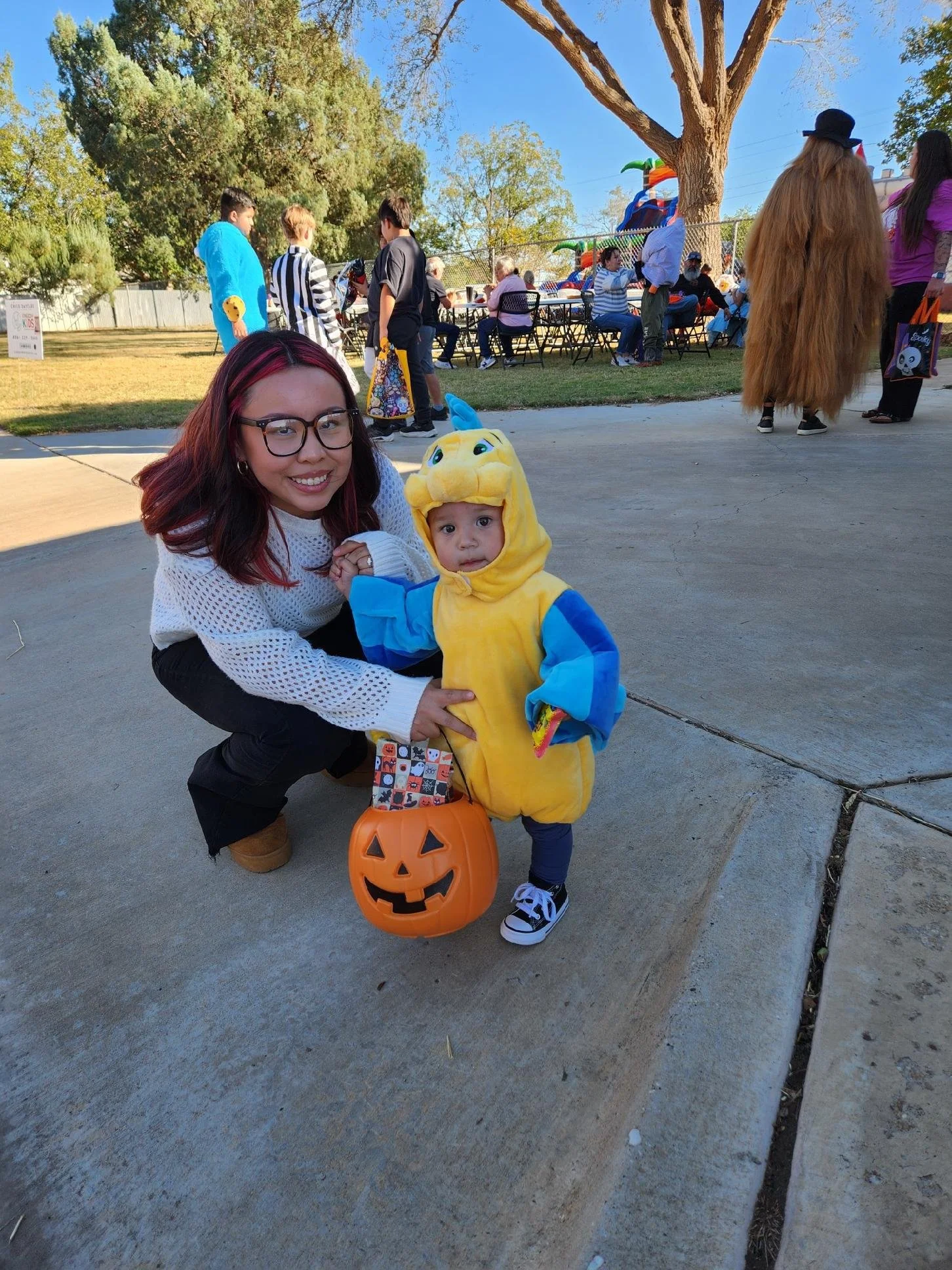 A woman with red-dyed hair and glasses crouches next to a young child dressed in a dragon costume at a Halloween event. The child holds a pumpkin-shaped basket with a carved face. In the background, other people are gathered outside, some in costumes
