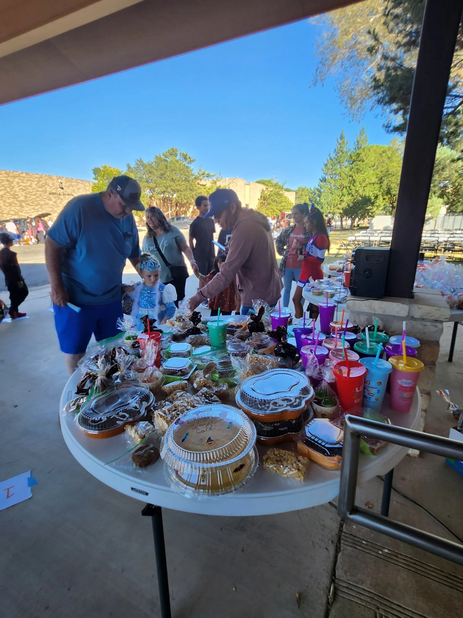 A table with assorted baked goods, cupcakes, candies, and colorful drinks at an outdoor gathering under a pavilion, with people in line and children nearby.