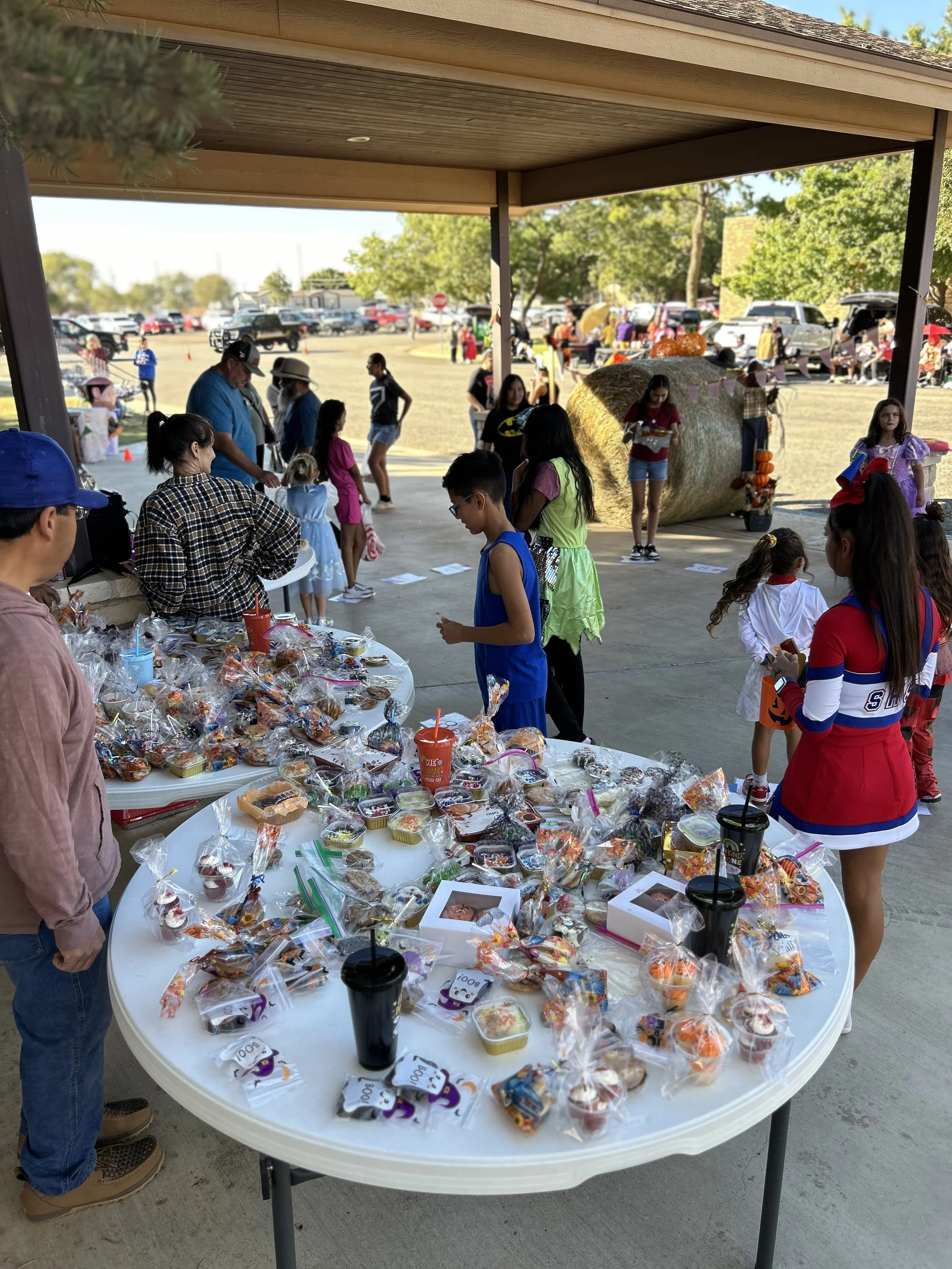Children and adults at a fall festival, with a table of Halloween treats and cups, a hay bale decoration, and pumpkins in the background.