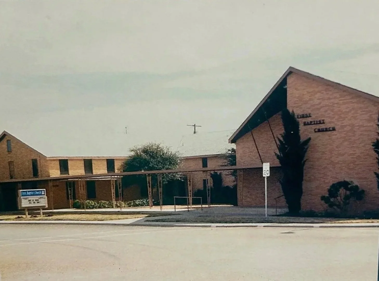 Exterior view of the church with brick walls and a sloped roof, labeled 'First Baptist Church,' with trees and a sidewalk in front.