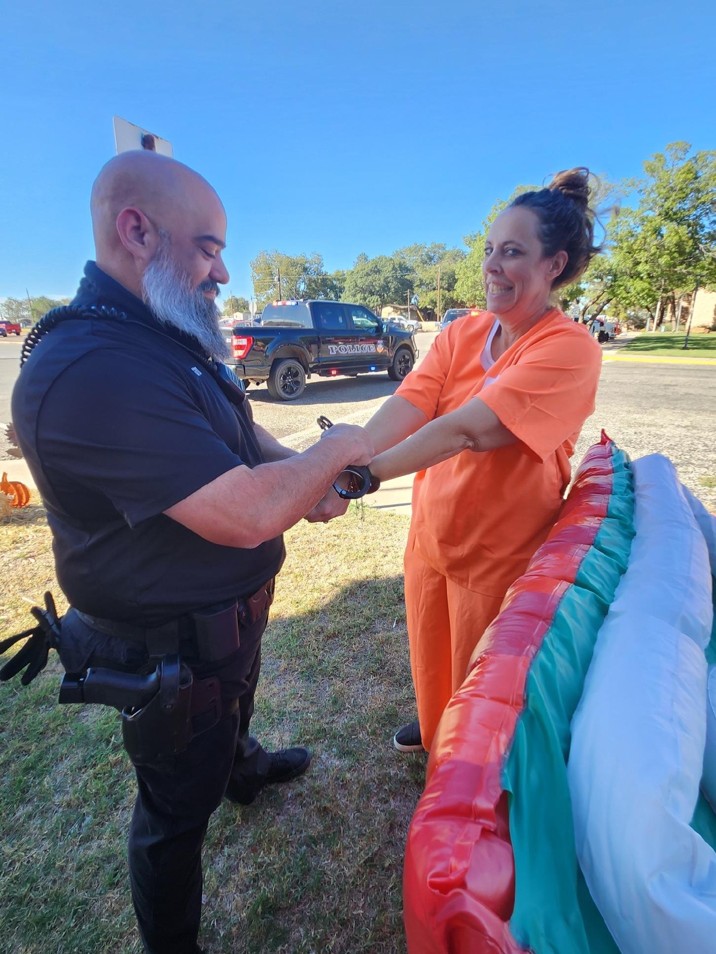 A police officer and a woman in orange scrubs are outside on a sunny day, holding hands and smiling. The police officer is on the left, with a bald head and gray beard, and the woman is on the right, with curly hair tied up. There are police cars and