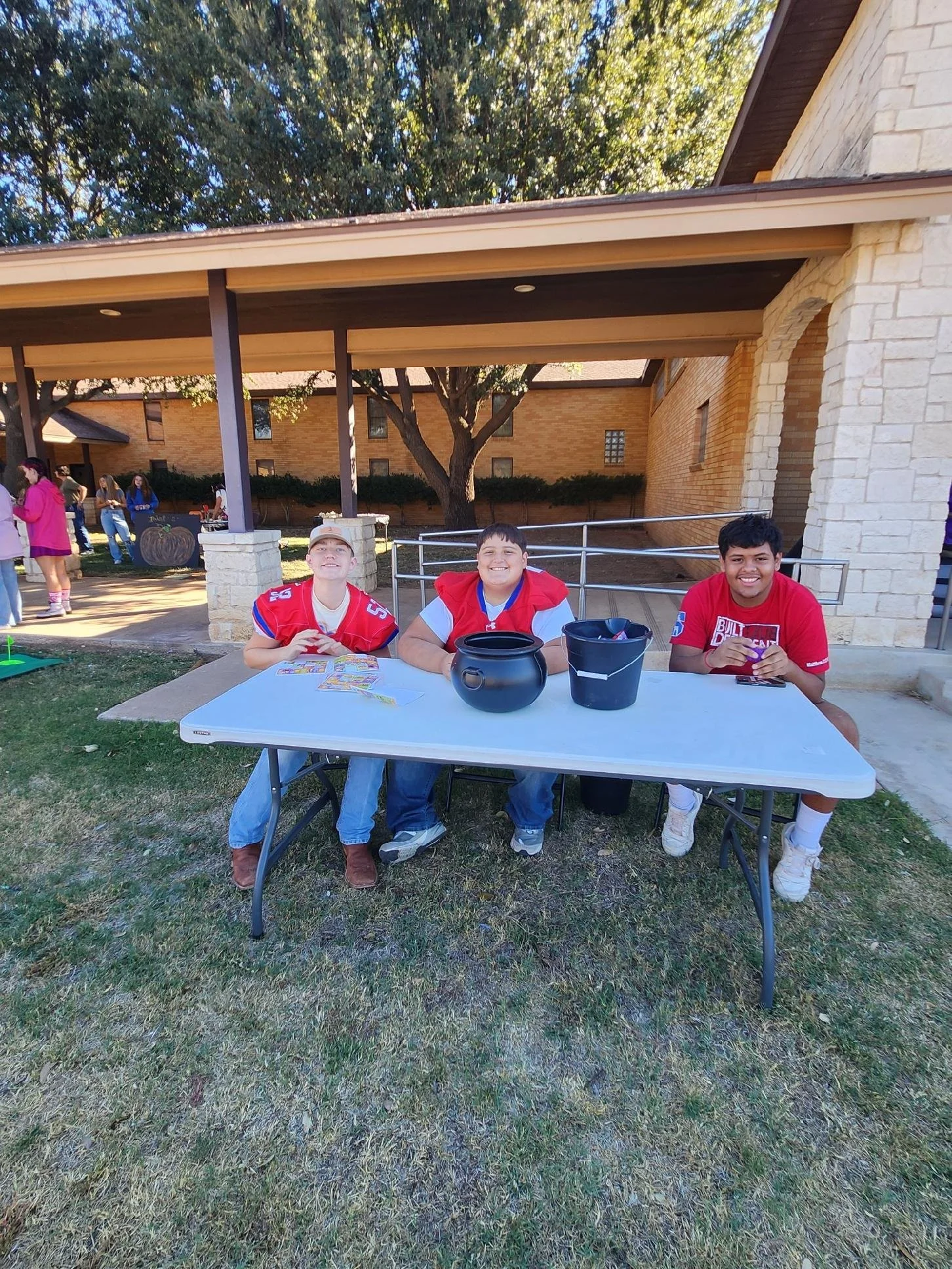 Three boys sitting at a table outdoors during daytime, wearing football jerseys, with buckets and cards on the table, smiling at the camera.