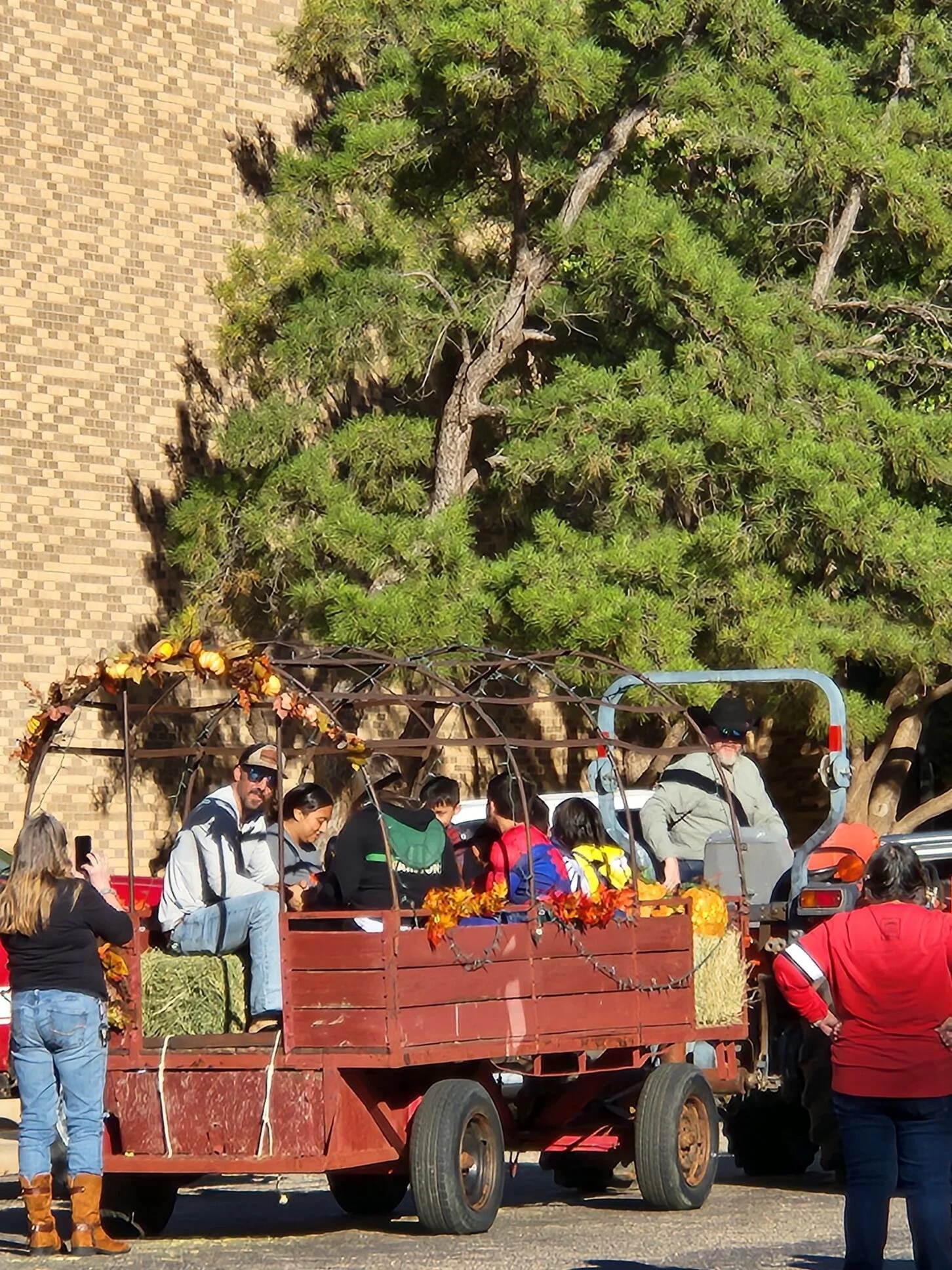 People riding on a decorated country wagon pulled by a tractor during a fall festival, with a backdrop of green trees and a brick wall.