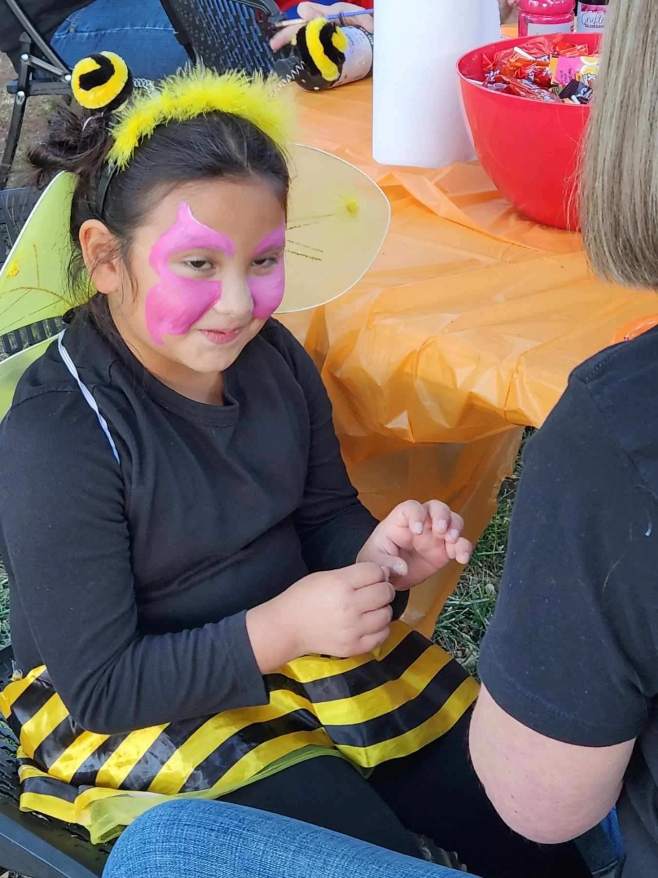 A young girl dressed as a bee, with face paint resembling a butterfly, wearing bee-themed accessories, sitting at a table with party supplies, and smiling at someone.