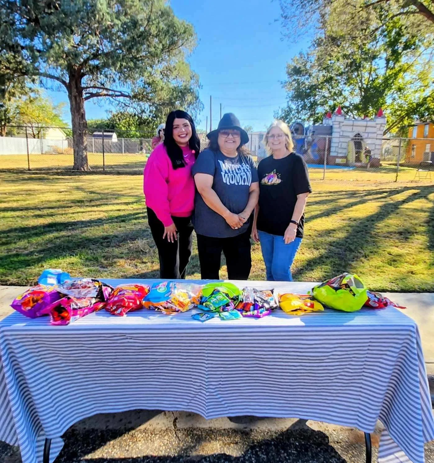 Three women standing behind a table with various snack bags, outdoors in a park with a fence, trees, and a bounce house in the background.