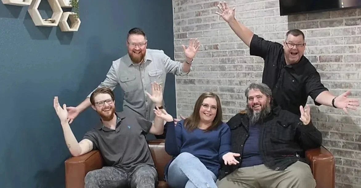Group of five people sitting and standing on a sofa, smiling, with two women and three men, in a casual indoor setting with a brick wall and a blue accent wall.