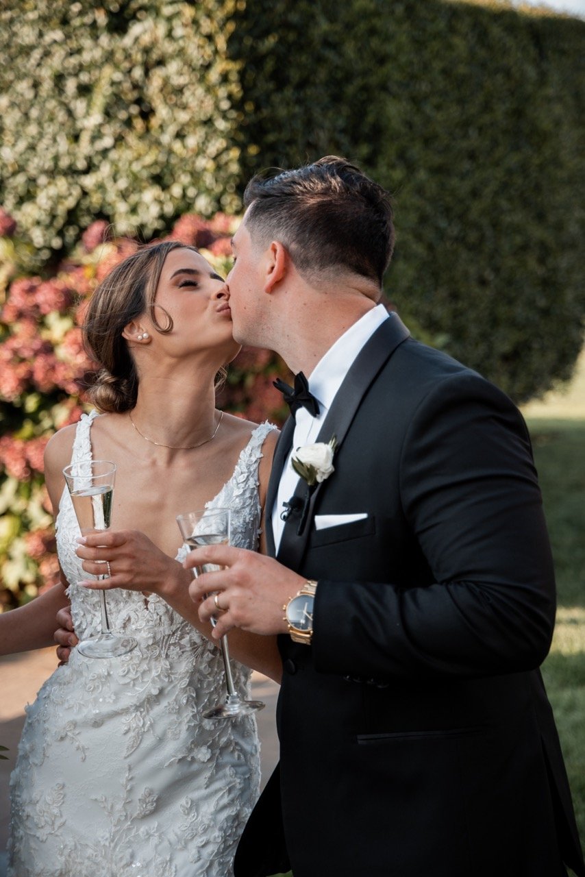 Bride and groom kissing at their wedding ceremony outdoors, holding champagne glasses, with a background of flowers and greenery.